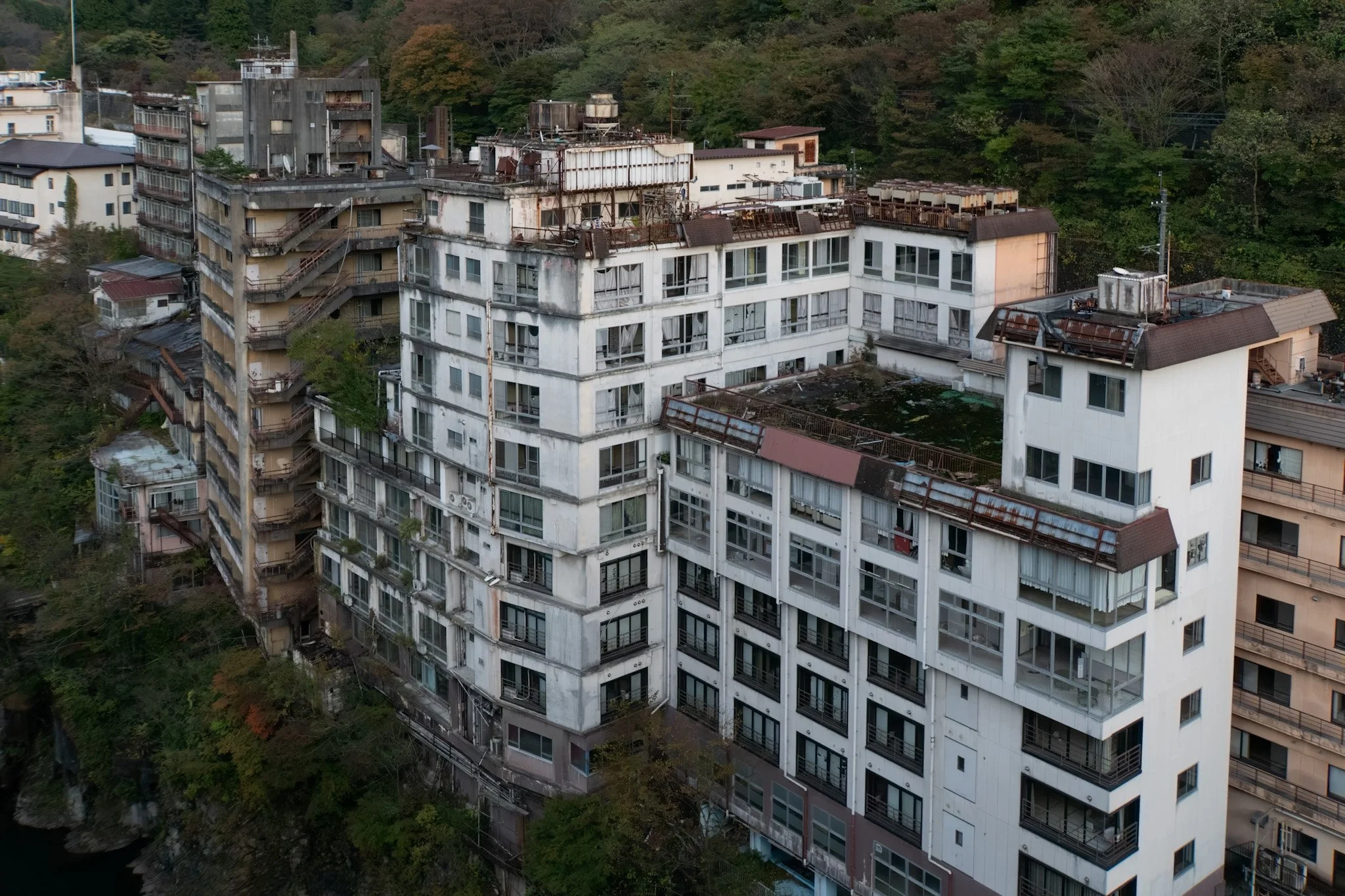 An aerial view of an old, multi-story apartment building with worn-out exterior walls, rusted railings, and broken or missing windows, set against a backdrop of green trees.