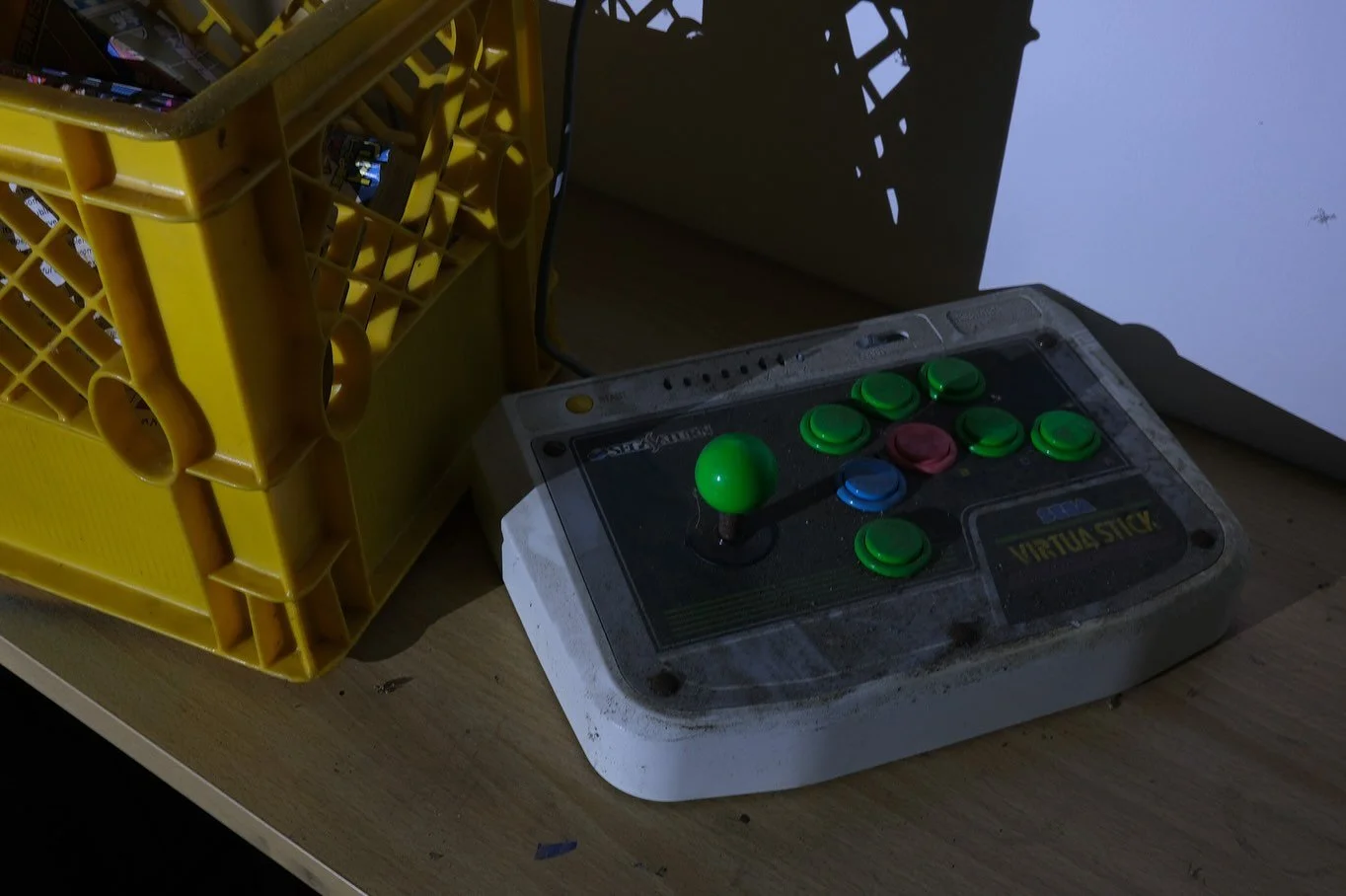 Dust-covered arcade controller with a green ball top, various green buttons, the words "Virtua Stick" on it, and a yellow plastic crate beside it, illuminated by flashlight in the installation. An arcade joystick controller 