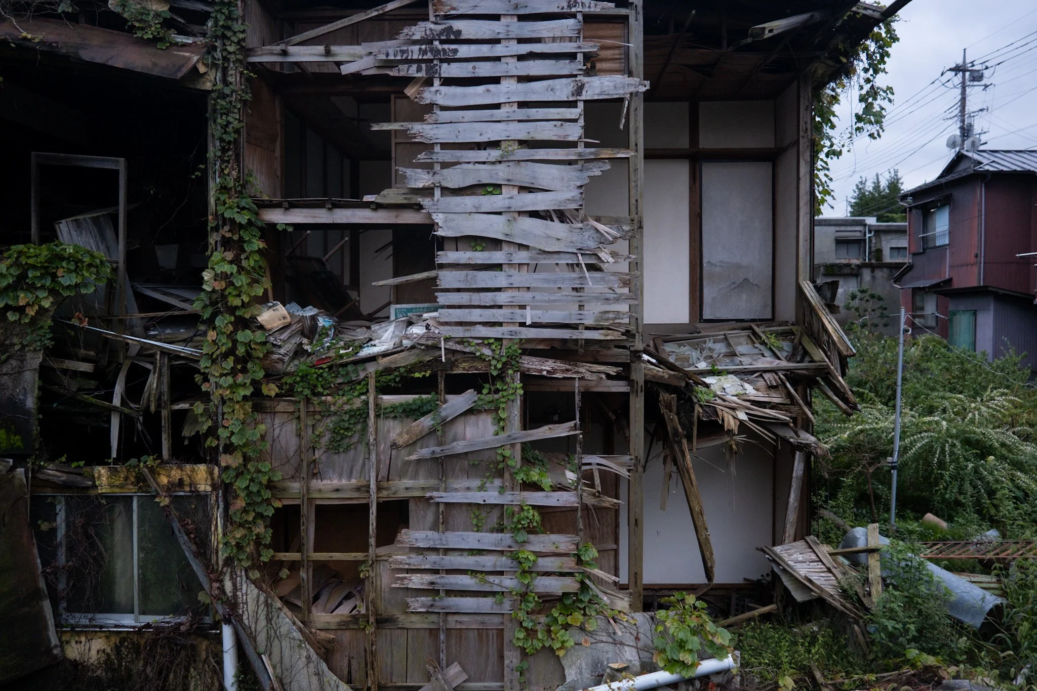 A partially collapsed wooden house with broken and missing planks, overgrown with vines and surrounded by other residential buildings and utility poles, in Kinugawa Onsen.