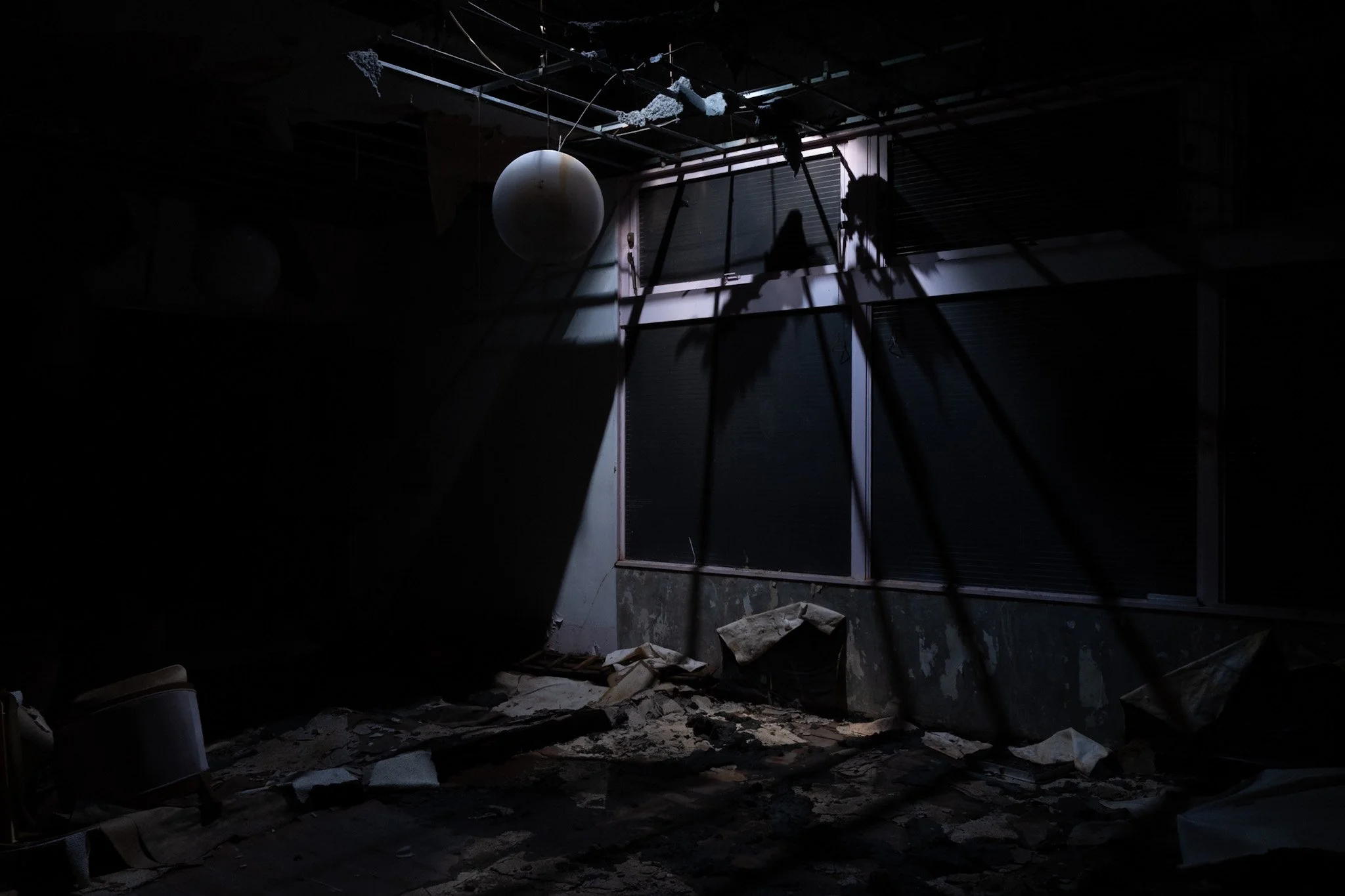 Ballroom of the abandoned Green Palace Hotel in Kinugawa Onsen, with broken ceiling, scattered debris on the floor, and a window with closed blinds. Shadows cast by the window frame and debris create a haunting atmosphere.