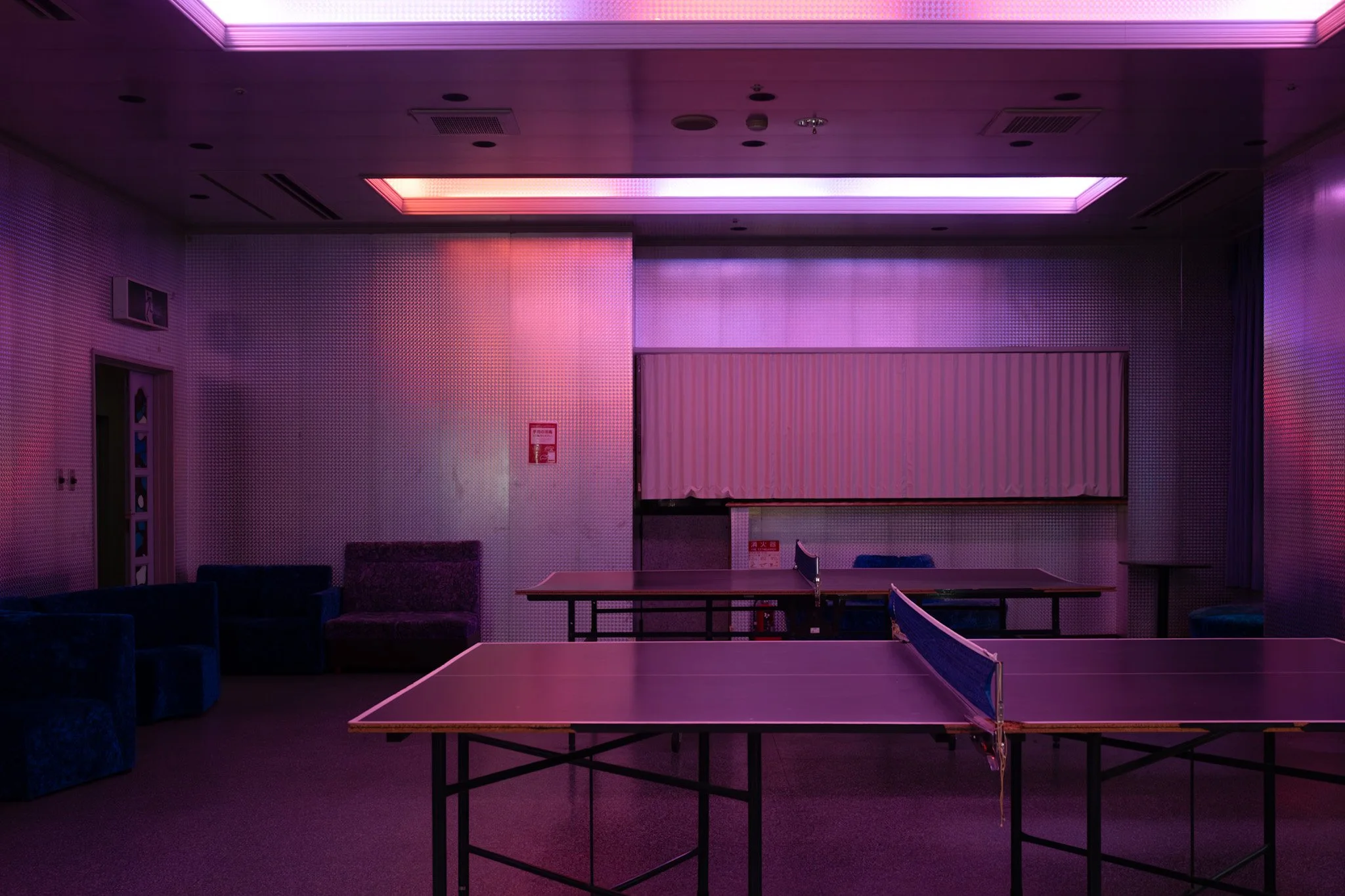 Empty hotel recreation room with ping-pong tables under purple and pink lighting.