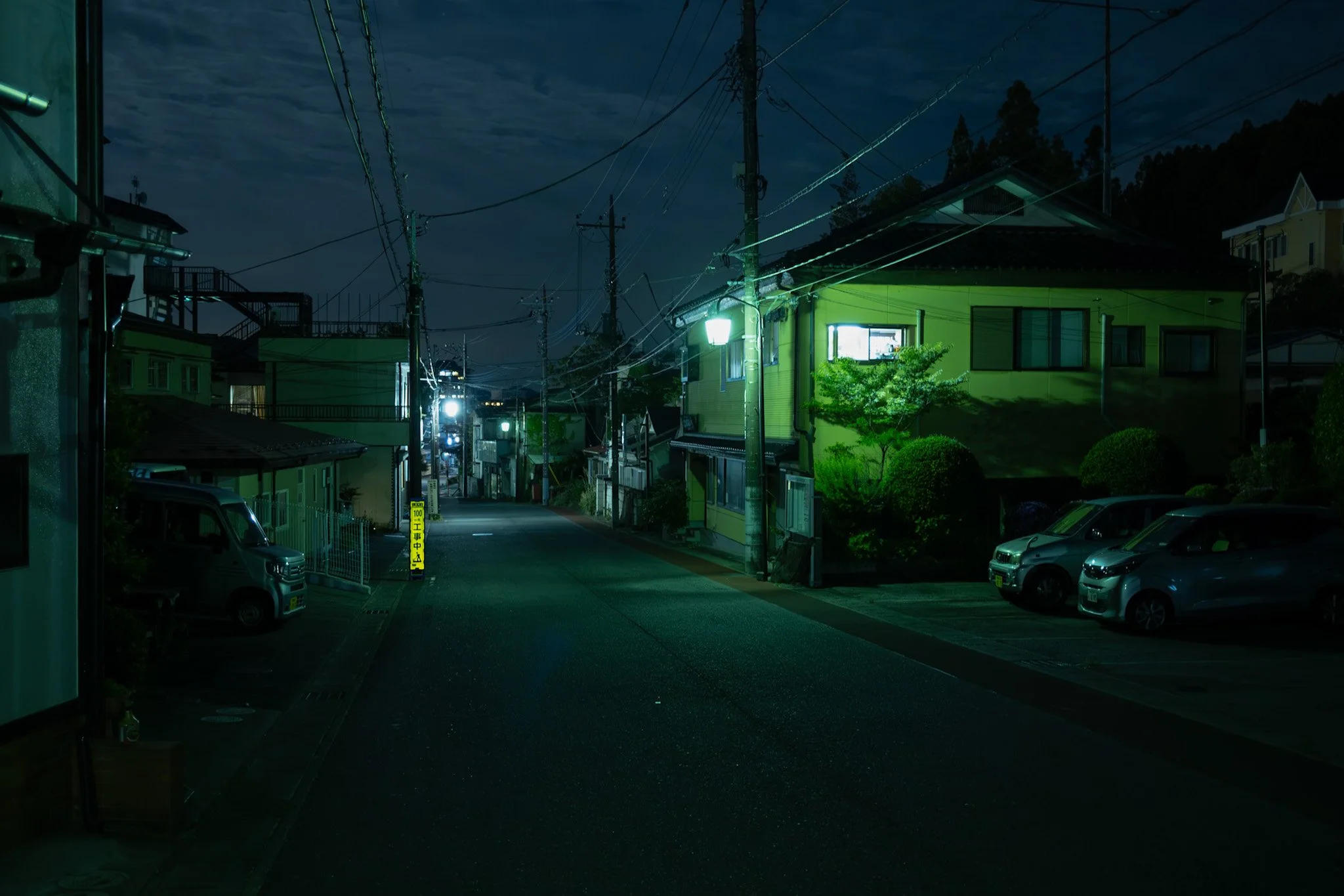 Nighttime view of a residential street in Kinugawa Onsen. With houses, parked cars, utility poles, and overhead power lines, illuminated by green streetlights and house lights, with a cloudy sky in the background.