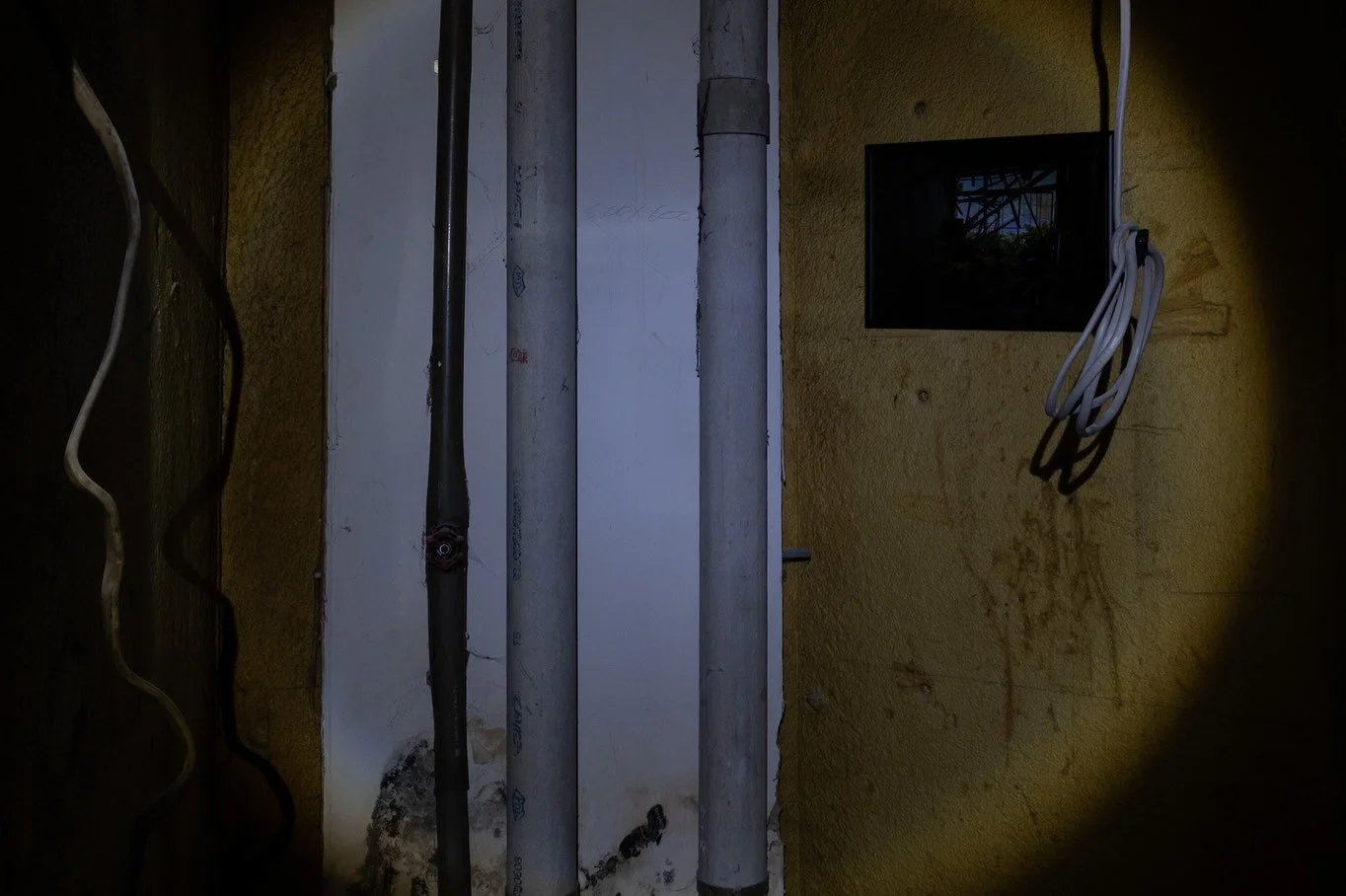 Close-up of an interior wall with electrical cables, pipes, and a framed photograph, illuminated by flashlight inside the Ghosts of Tomorrow installation.