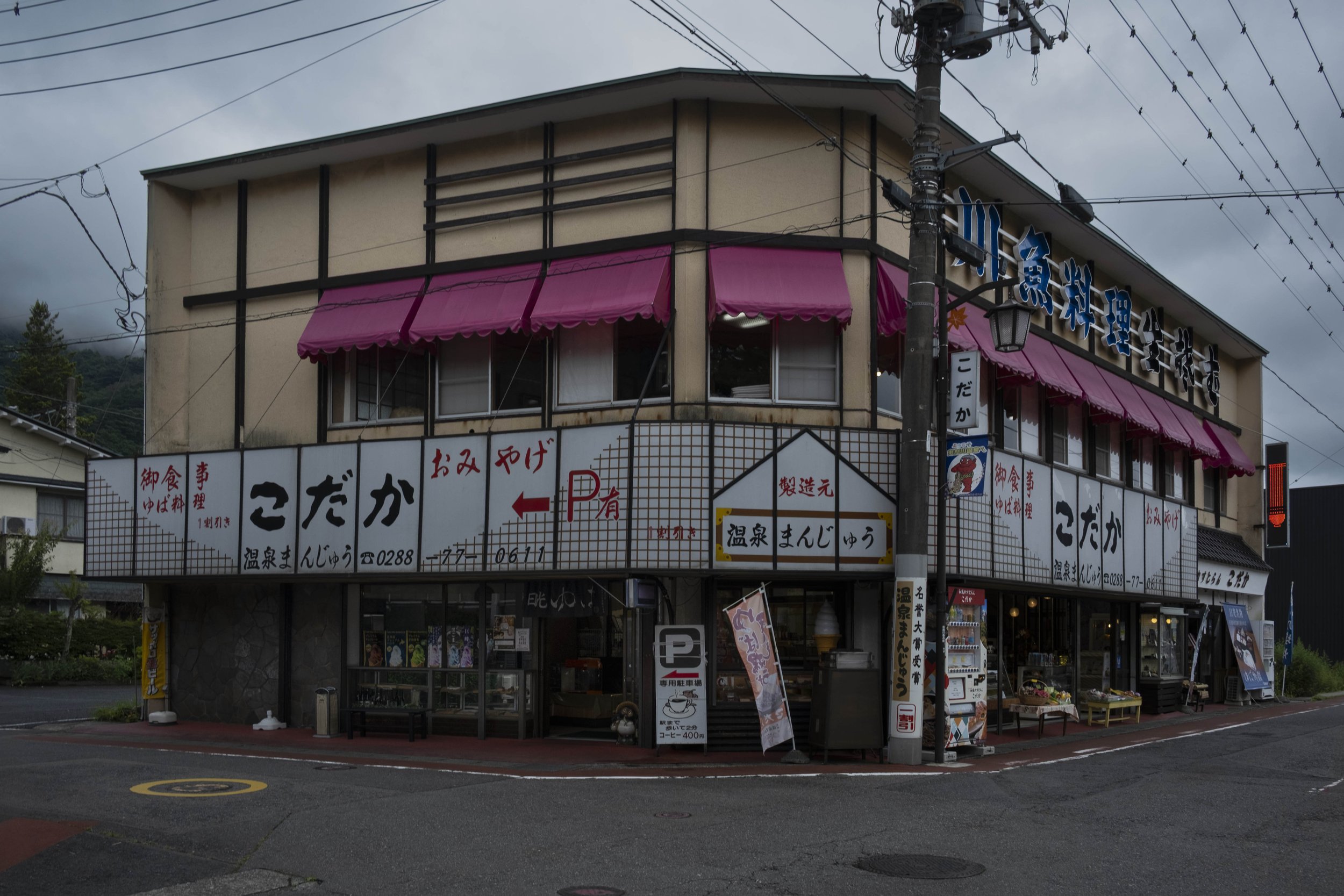 Exterior of Kodaka souvenir and food shop in Kinugawa Onsen. A two-story building with signs in Japanese, pink awnings over the windows, and various signs and advertisements on the lower level. There is a utility pole with wires in front of the build