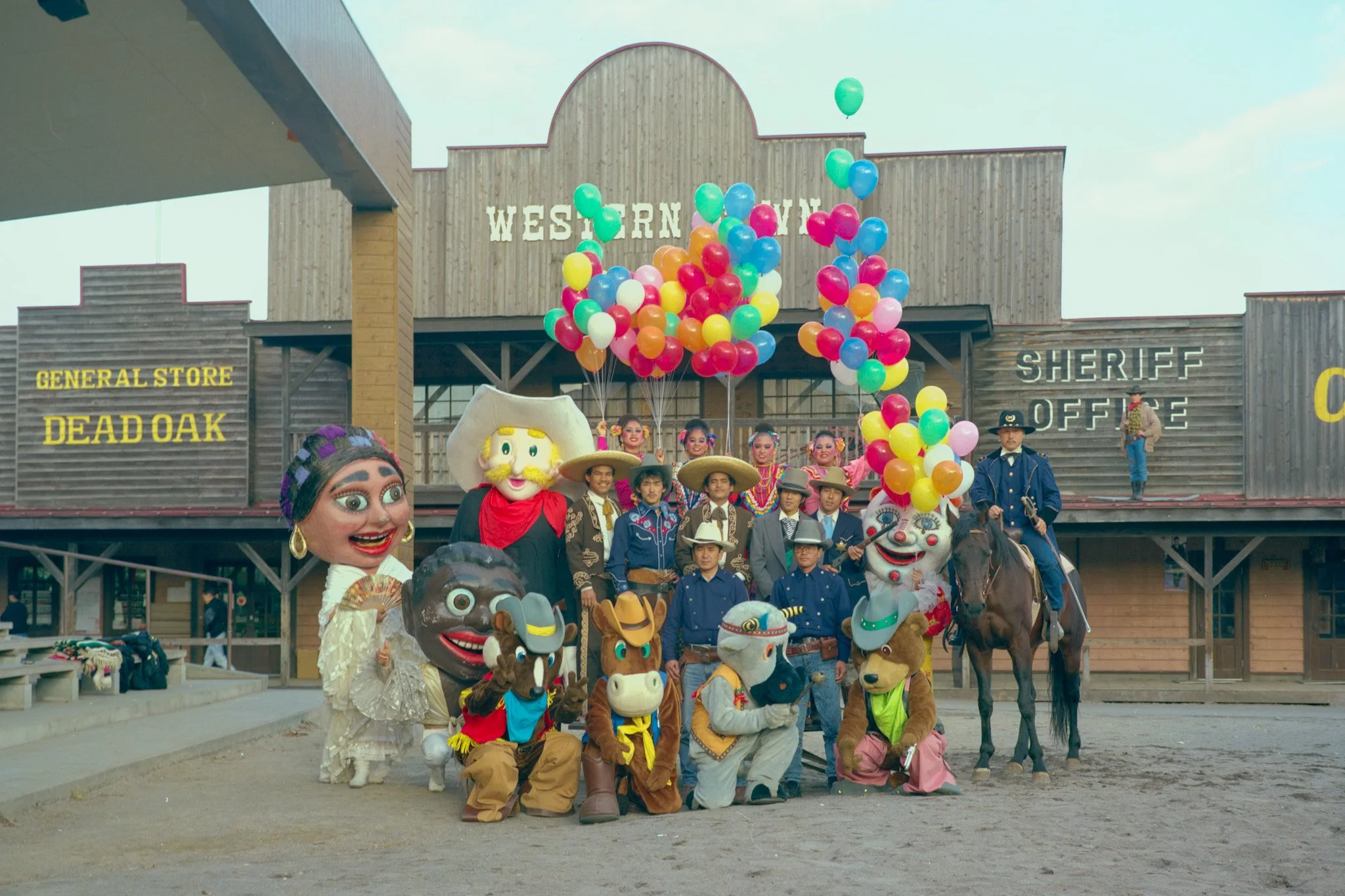 Found photograph of Western Village staff and performers posing. The scene features colorful balloons, some mounted on horseback and others floating in the air, with a bright, clear sky above.