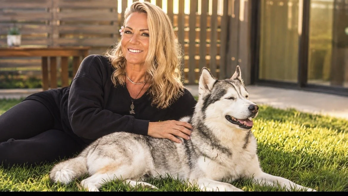 A woman lying on grass next to a smiling Siberian Husky dog with a partly sunlit backyard in the background.
