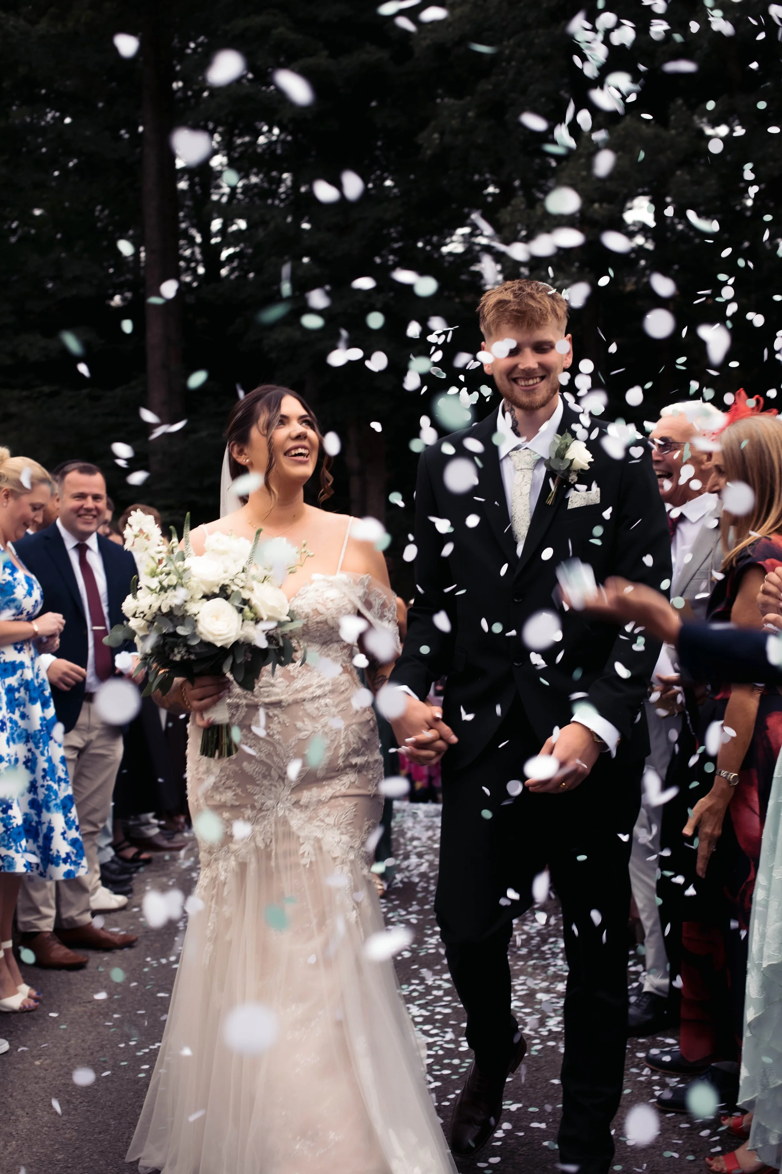 A bride and groom smiling and holding hands, surrounded by guests throwing white confetti at an outdoor wedding celebration.