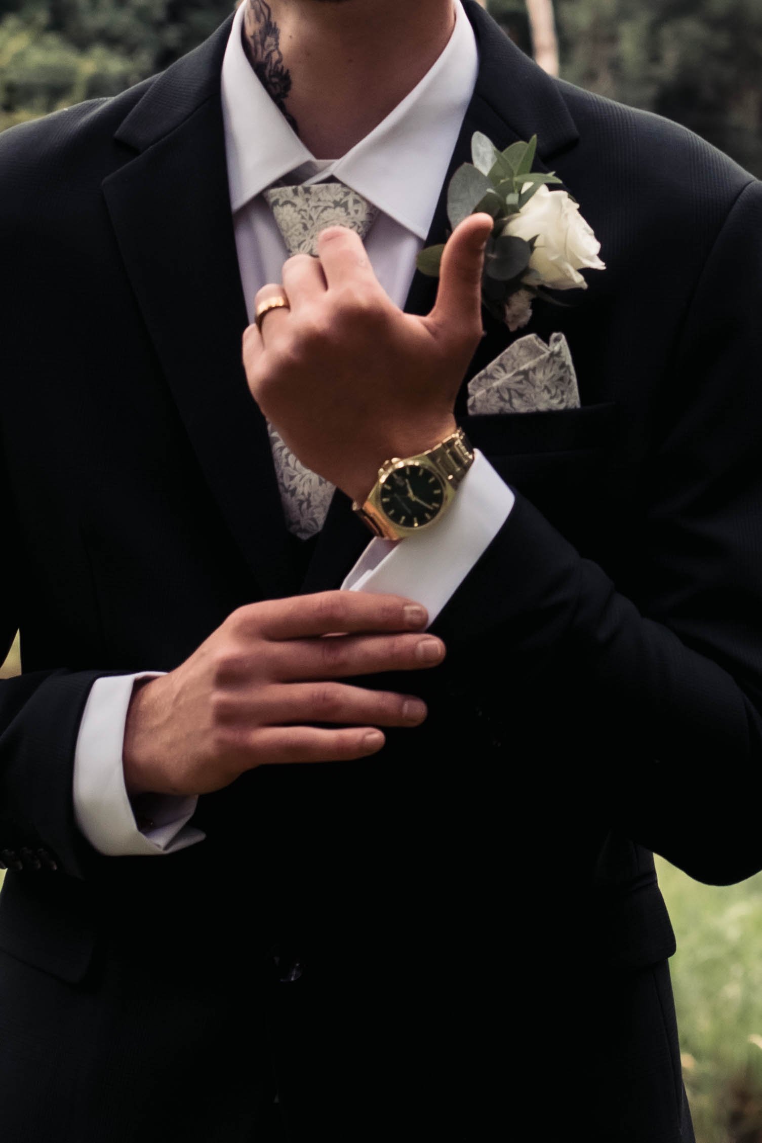 Close-up of a man in a tuxedo adjusting his shirt with a boutonniere pinned to his lapel, wearing a gold watch and ring, set outdoors.