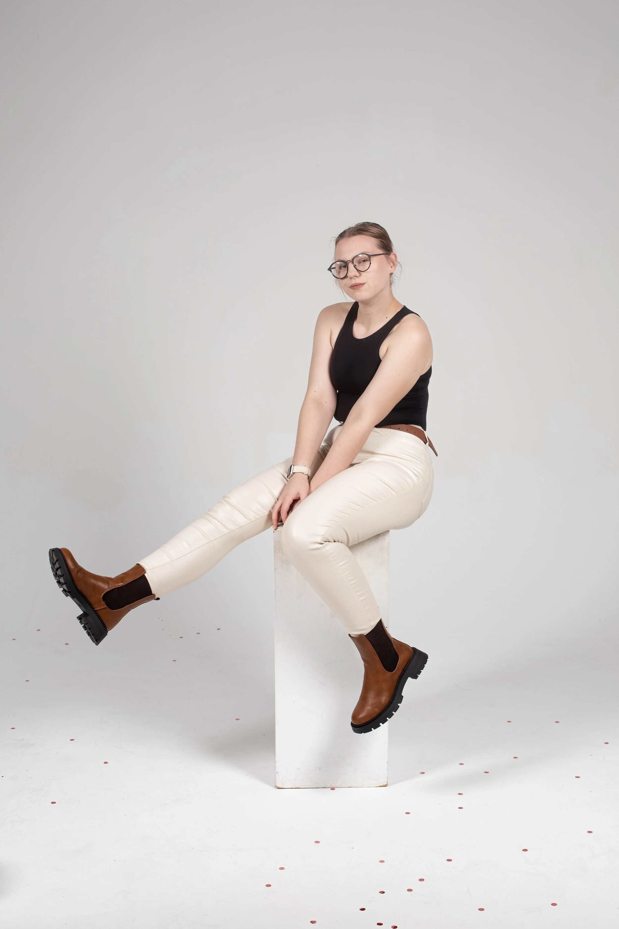 Young woman with glasses wearing a black tank top and cream pants, sitting on a white block against a plain gray wall, with her left leg extended and shoes resting on the floor.