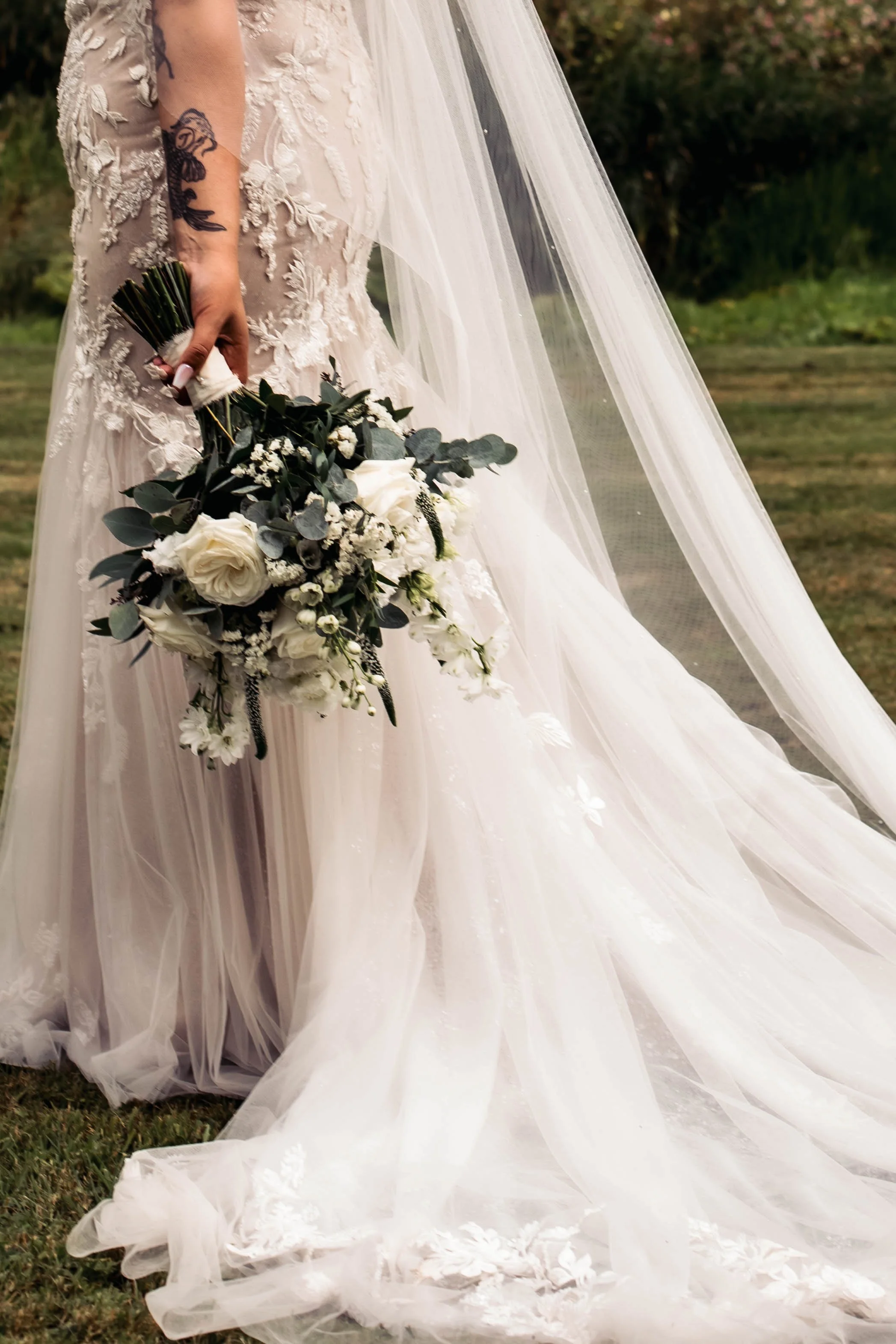 Close-up of a bride holding a bouquet of white roses, greenery, small white flowers, and black accents, wearing a wedding dress with lace details and a long tulle veil, outdoors on grass with bushes in background.