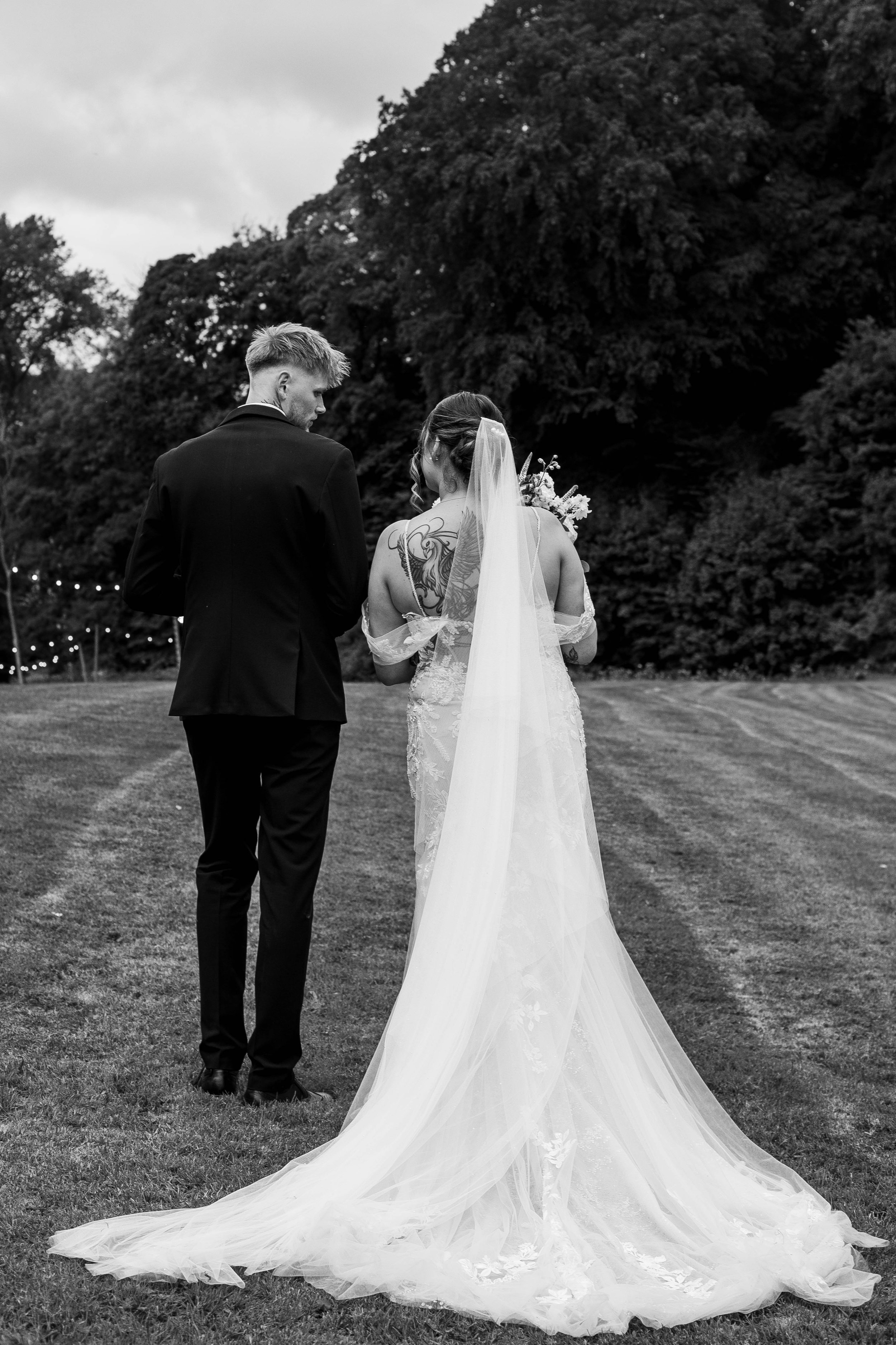 Black and white photo of a bride and groom outdoors, standing on grass with trees in the background. The groom is wearing a suit and the bride a wedding gown with a long train, and she has a veil and a tattoo on her shoulder. They are facing each other, appearing to share an intimate moment.