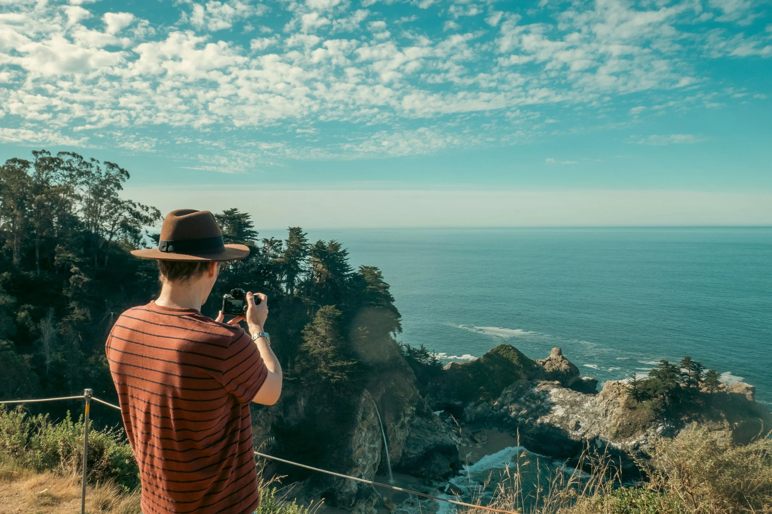 A person wearing a brown hat and a red striped shirt takes a photo with a camera during daytime, overlooking a scenic coastal landscape with trees, rocky cliffs, and the ocean under a partly cloudy sky.