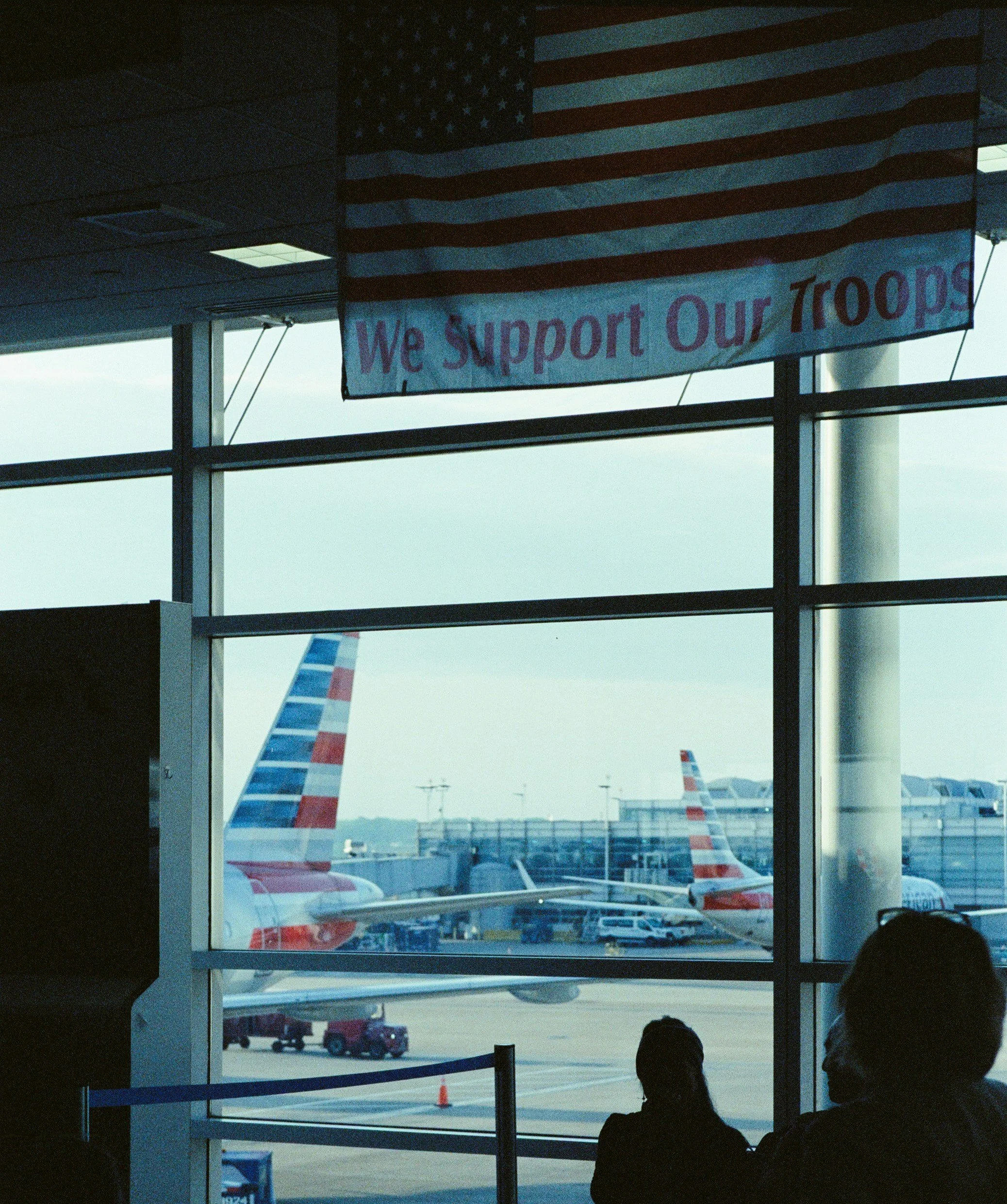 American flag and a banner supporting troops inside an airport terminal with airplanes visible through the windows.