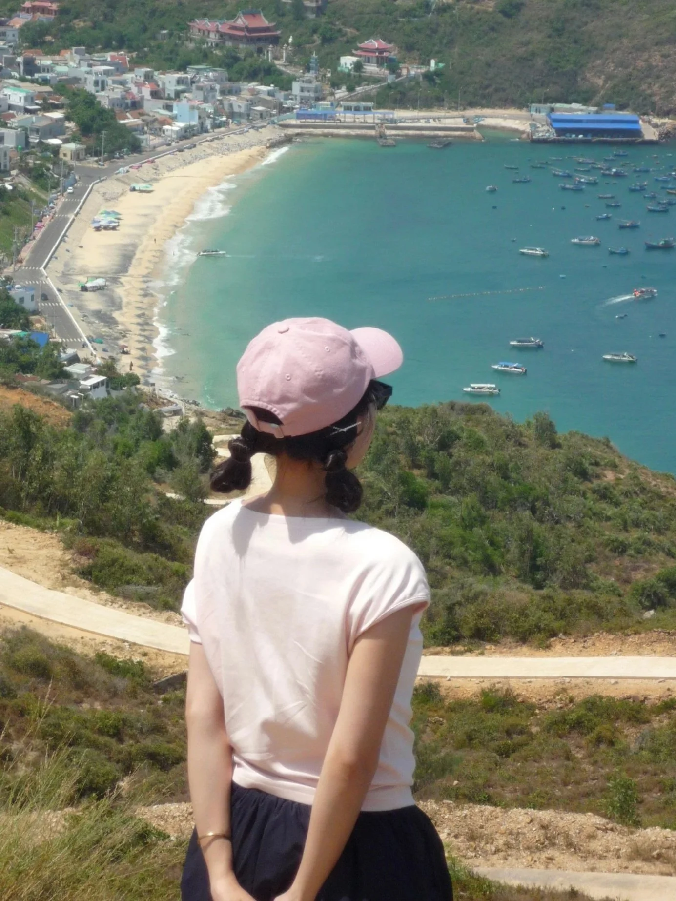 A woman with braided hair in a pink cap and white shirt standing on a hill overlooking a coastal town with a beach and numerous boats in the water.