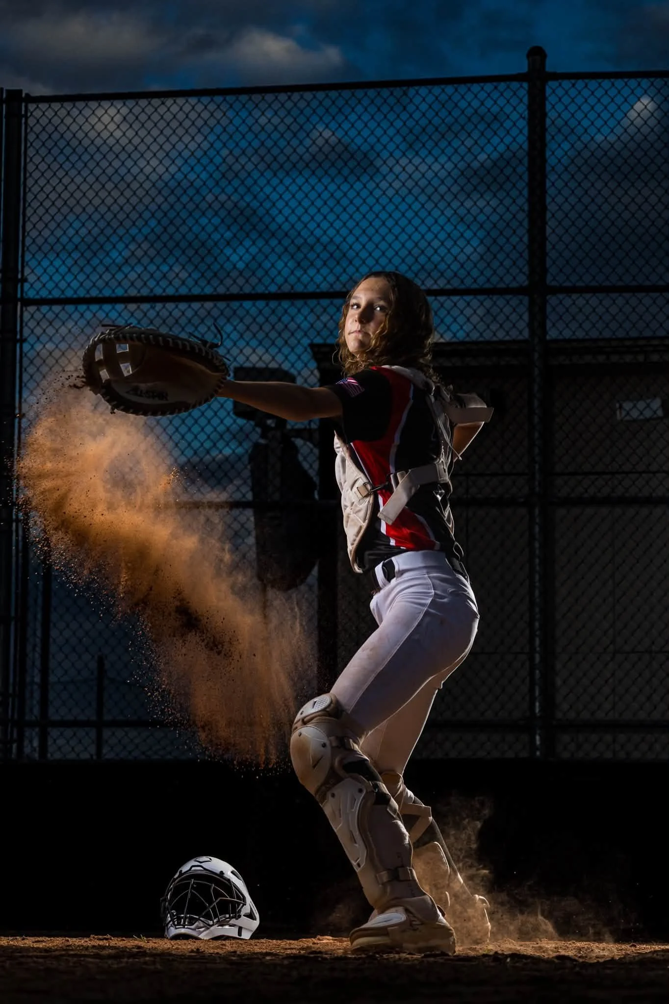 A female softball pitcher in a black, red, and white uniform pitching a ball. Her glove is in her left hand, and her right arm is extended after releasing the ball. Dust is flying up from the ground as she winds up her pitch. A catcher's mask and hel