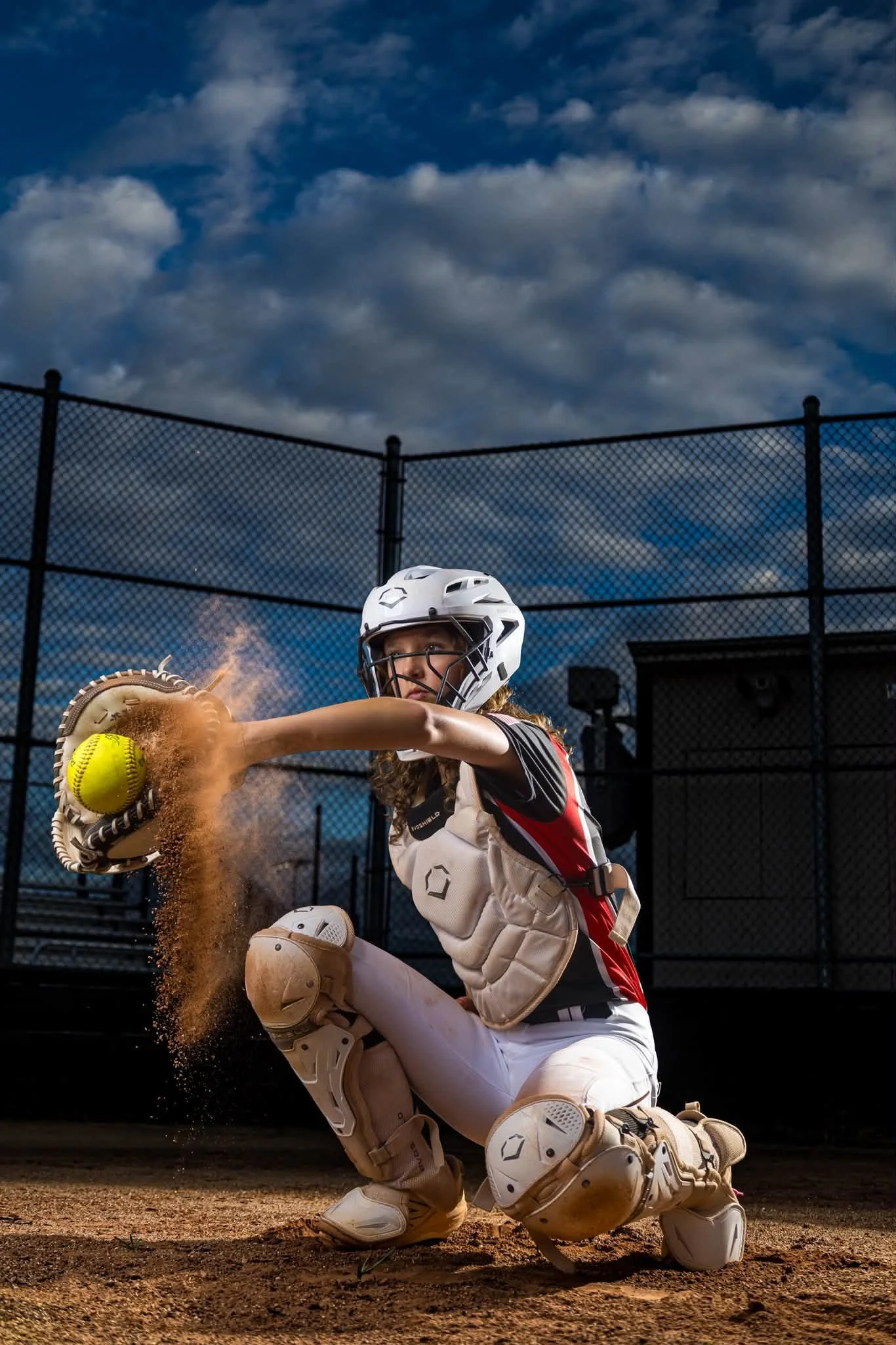 A female softball catcher in full gear making a tag during a game, with a dark sky and chain-link fence in the background.