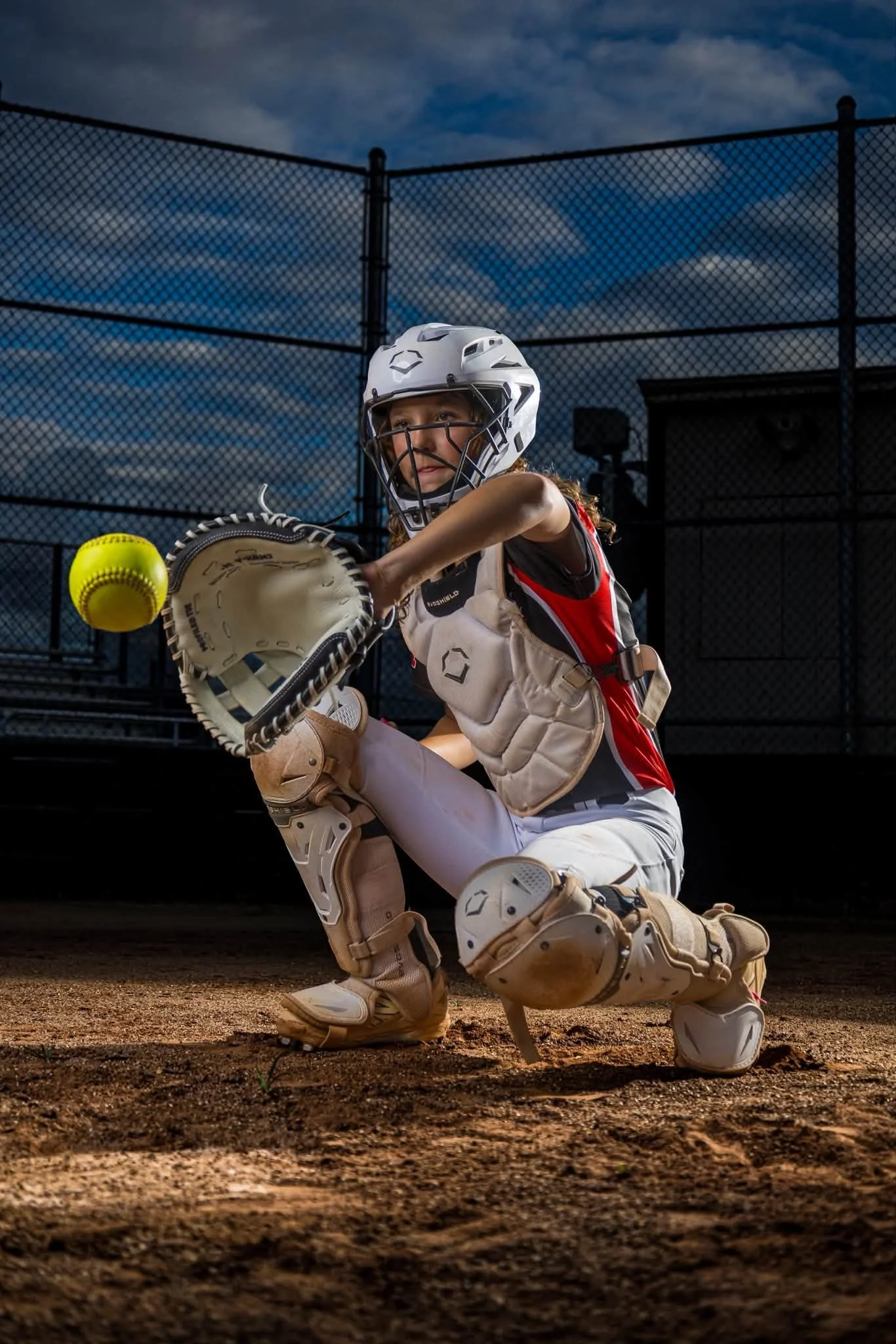 A baseball catcher wearing a helmet and protective gear, crouching behind home plate and preparing to catch a bright yellow baseball against a cloudy sky and a chain-link fence.
