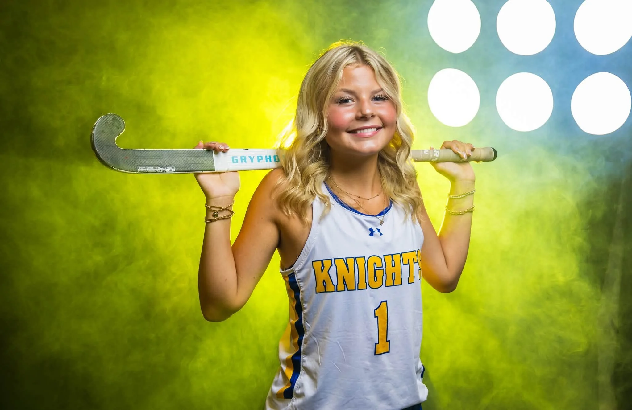A young girl with blonde curly hair smiling, wearing a white sports jersey with blue and yellow accents that says 'Knights', holding a hockey stick over her shoulders, against a colorful background with blue, green, and yellow hues and large white ci