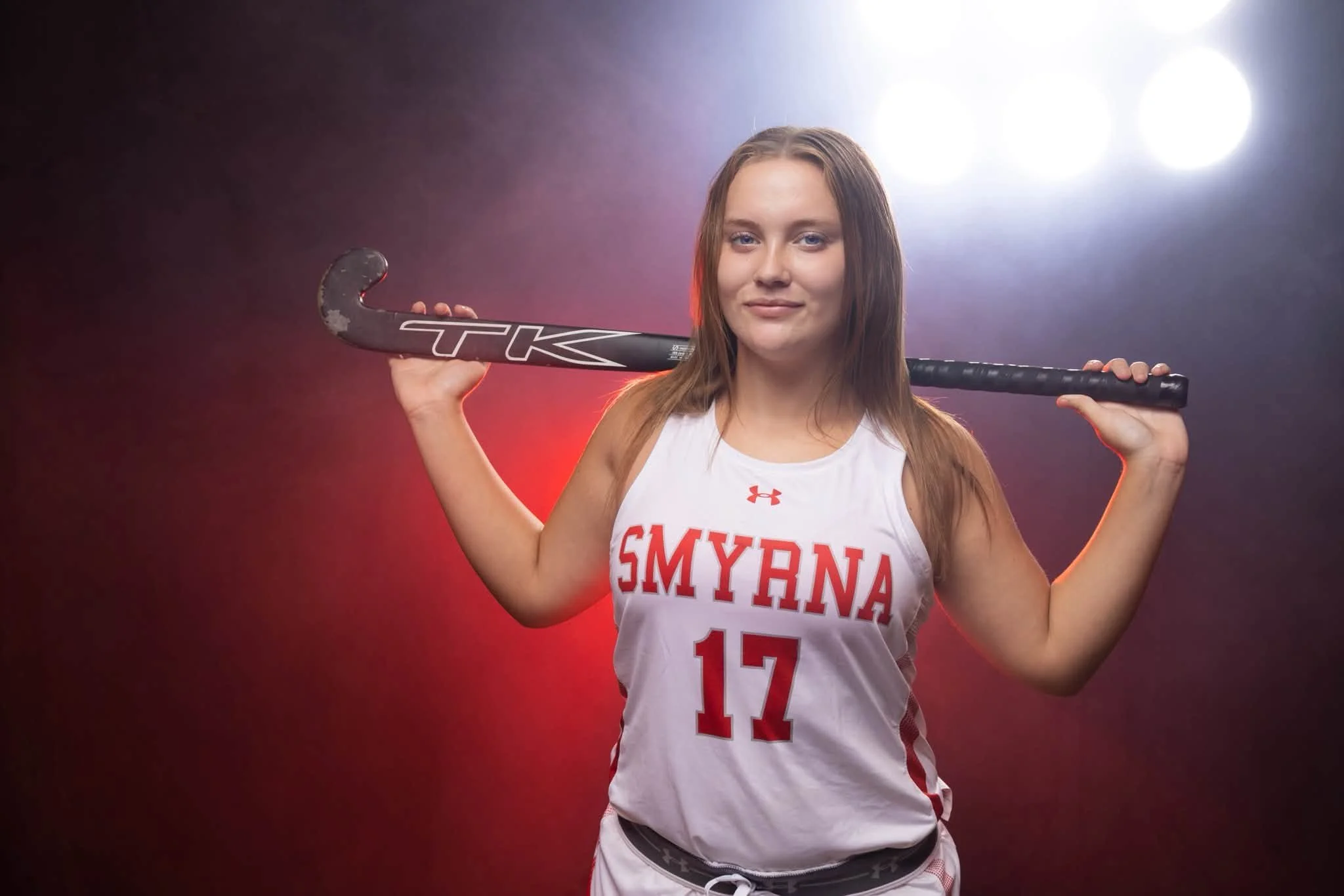 Young female athlete with long brown hair, dressed in a white sports uniform with red lettering 'SMyRNA' and the number 17, holding a black hockey stick over her shoulders, standing against a studio background with bright lights.
