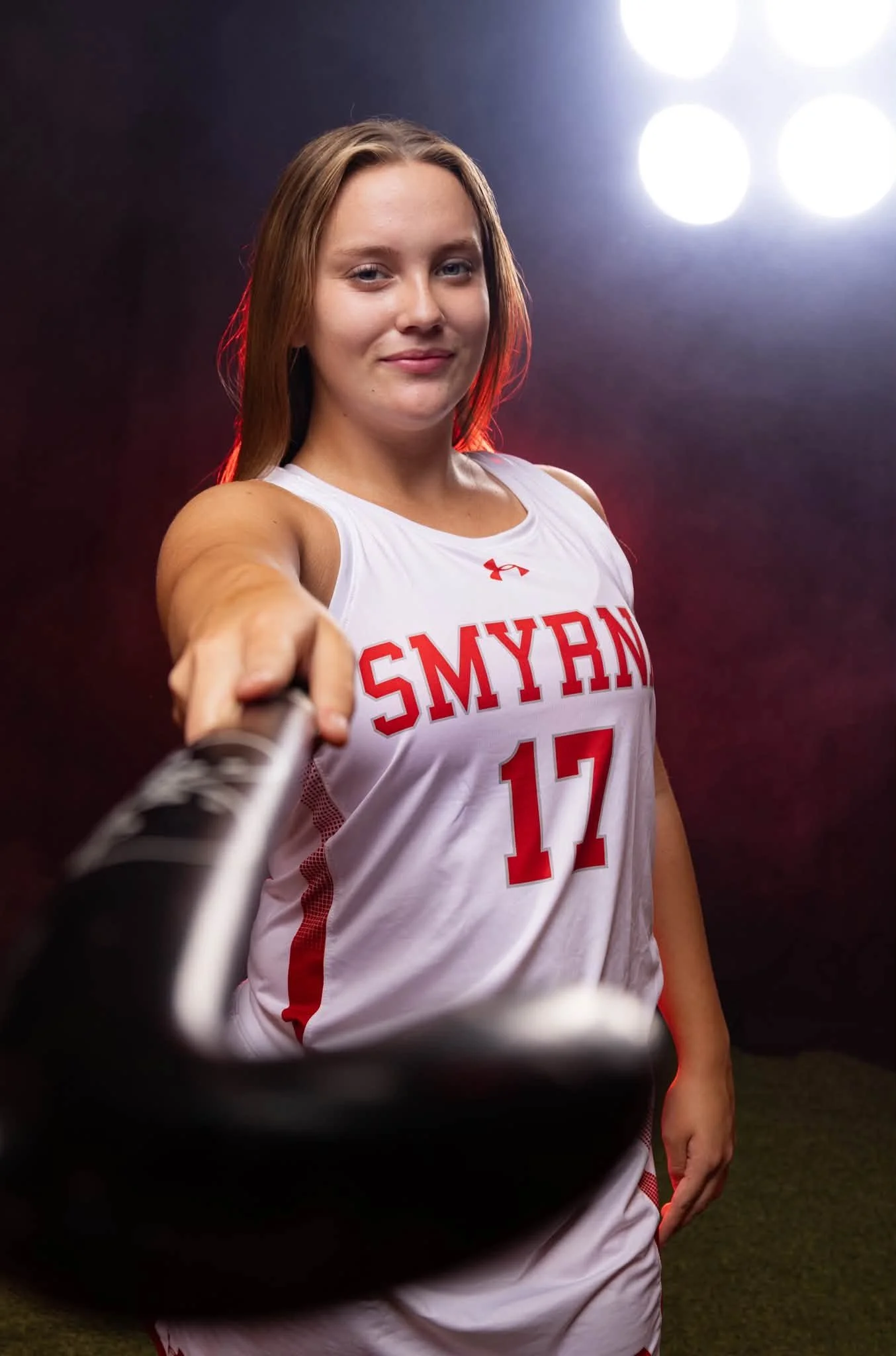 A young woman with long brown hair, wearing a white sports jersey with red lettering and the number 17, smiling and taking a selfie with the camera in front of her. Bright lights are visible behind her.
