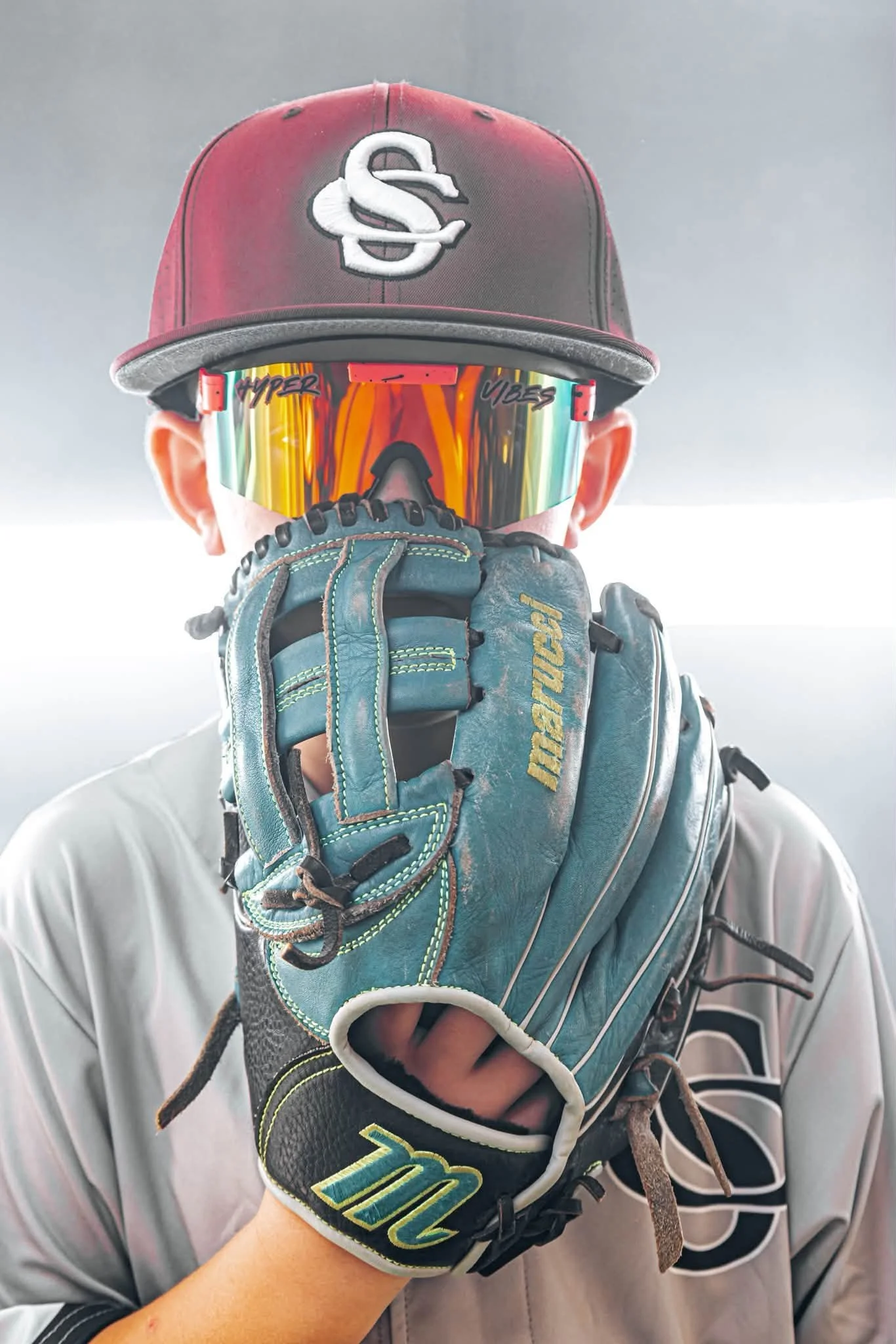 Person wearing a black baseball cap with white initials, mirrored sunglasses, and a baseball glove on their left hand, covering their face.