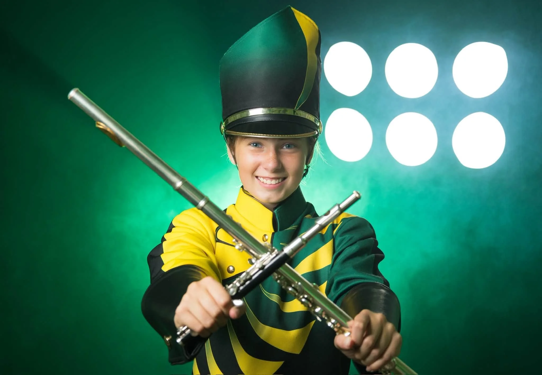 A young person dressed as a marching band member with a tall black hat and a yellow and green uniform, smiling while holding a silver flute against a green background with bright lights.