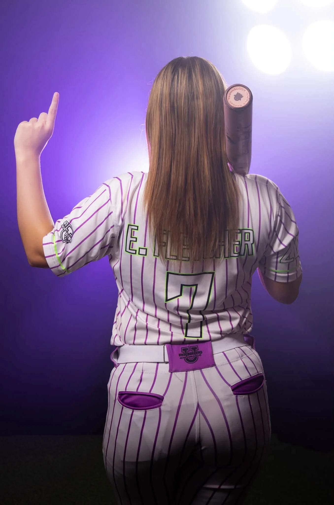 Back view of a female baseball player with long brown hair, wearing a white and purple pinstripe jersey with the number 7, and kneeling on the field with stadium lights in the background.