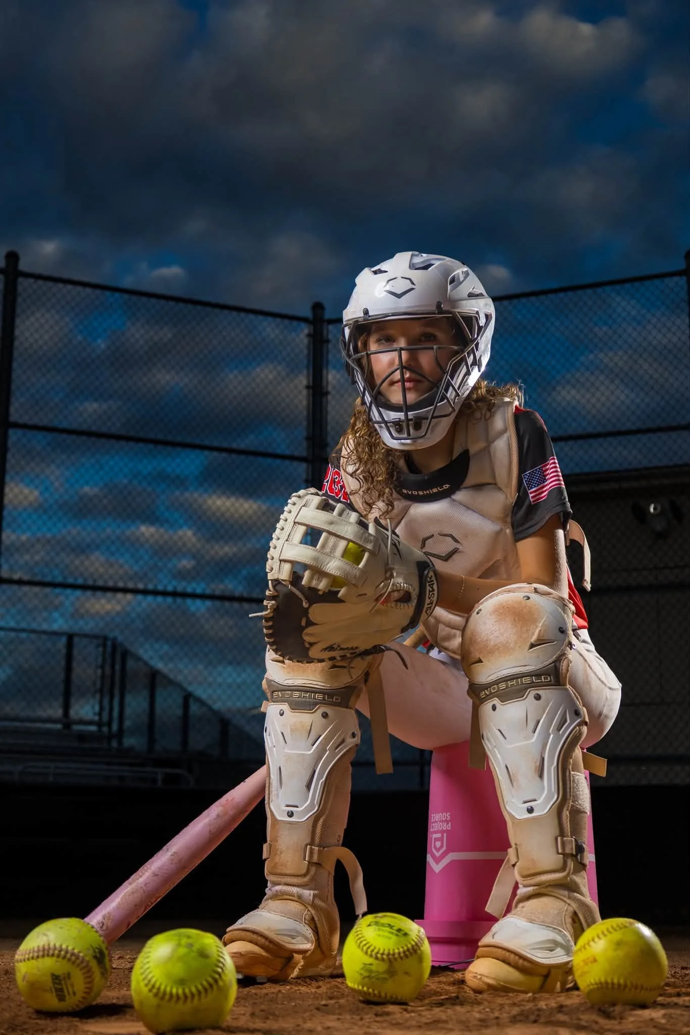 A young girl dressed as a softball catcher with a helmet and sports gear, kneeling behind the home plate, surrounded by yellow softballs, on a softball field with a chain-link fence and cloudy sky in the background.
