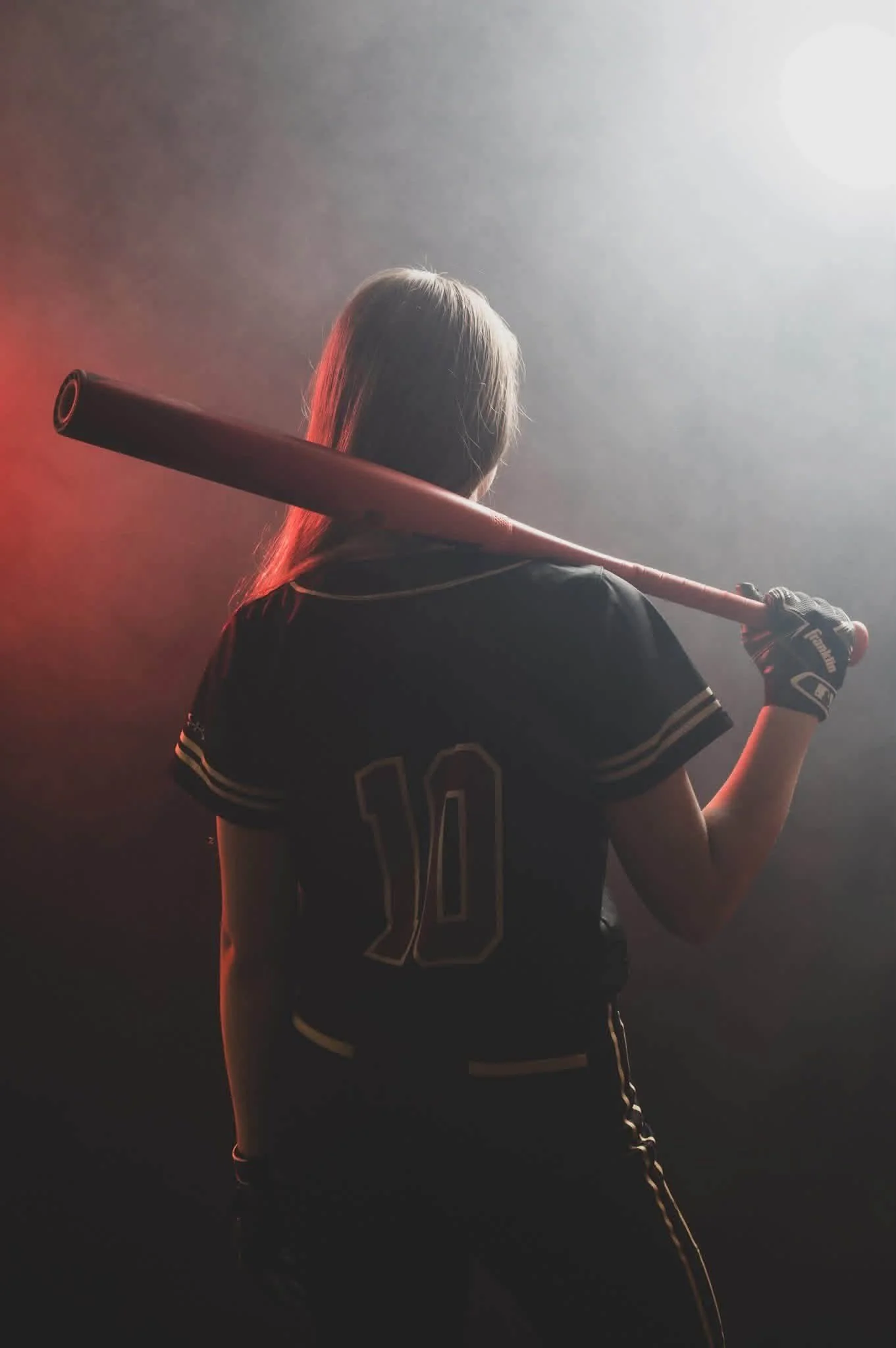 A female baseball player with blonde hair, wearing a black jersey with yellow trim and the number 10, holds a baseball bat over her shoulder in a hazy, dimly lit setting.