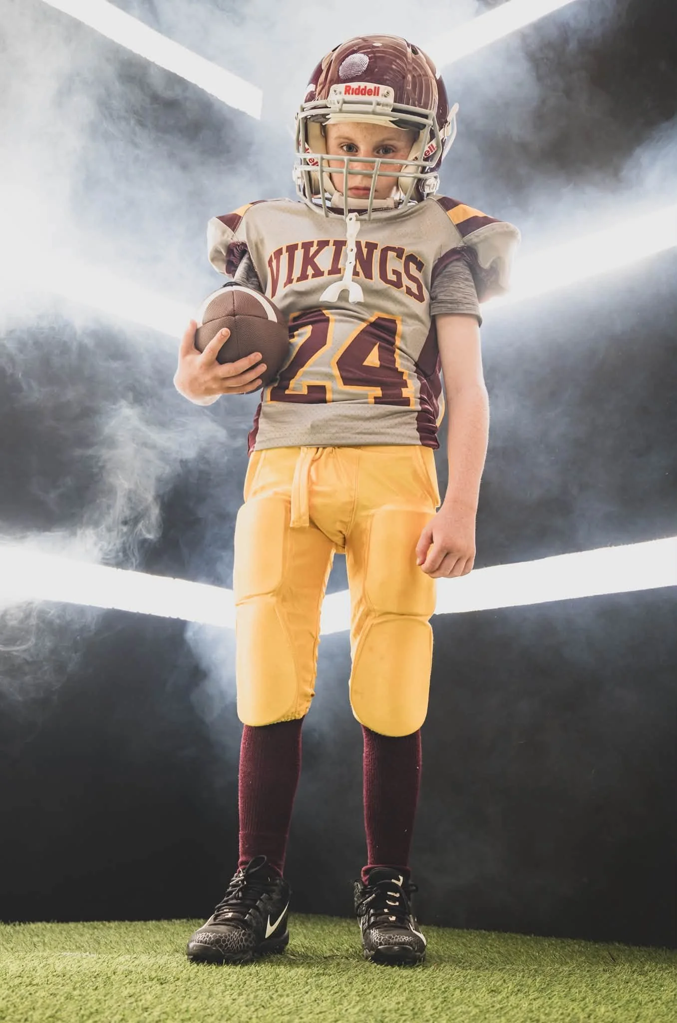 A young boy dressed in American football gear, holding a football and standing on a green turf field with a foggy or smoky background and bright lights overhead.