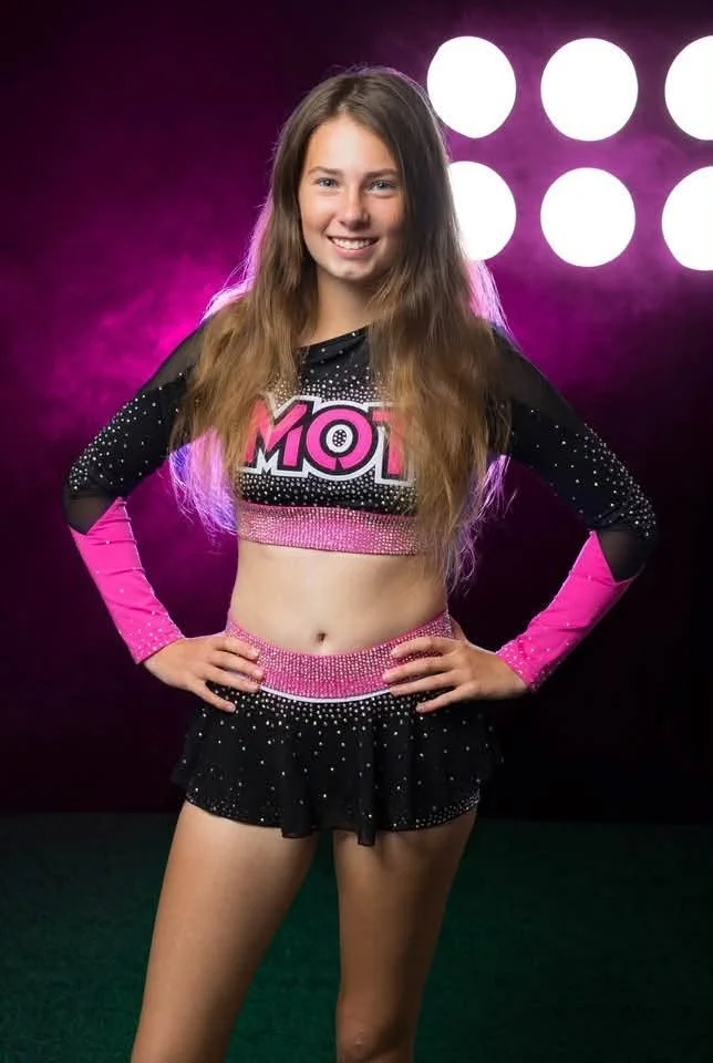 Teen girl in cheerleading outfit standing with hands on hips, smiling, pink and black sports uniform with rhinestones, background with purple lighting and four circular lights.