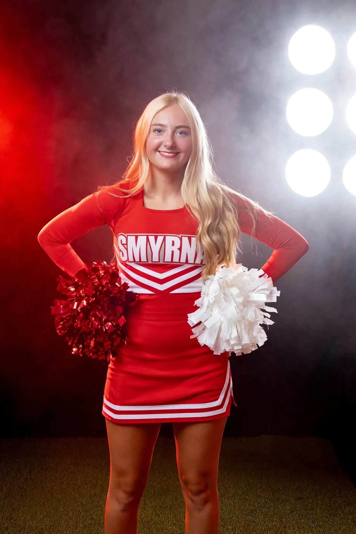 A young woman dressed as a cheerleader, wearing a red and white uniform with the word SMYRNA on the front, holding red pom-poms in her right hand and white pom-poms in her left hand, smiling at the camera, standing on a dark floor with bright lights 