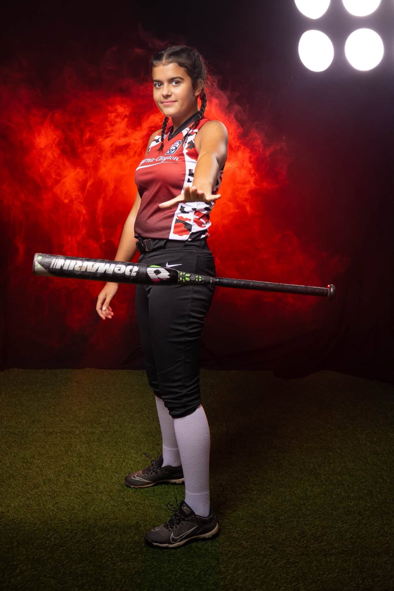 A young female athlete with braided hair holding a baseball bat inside a sports facility with a background of fire and bright lights.