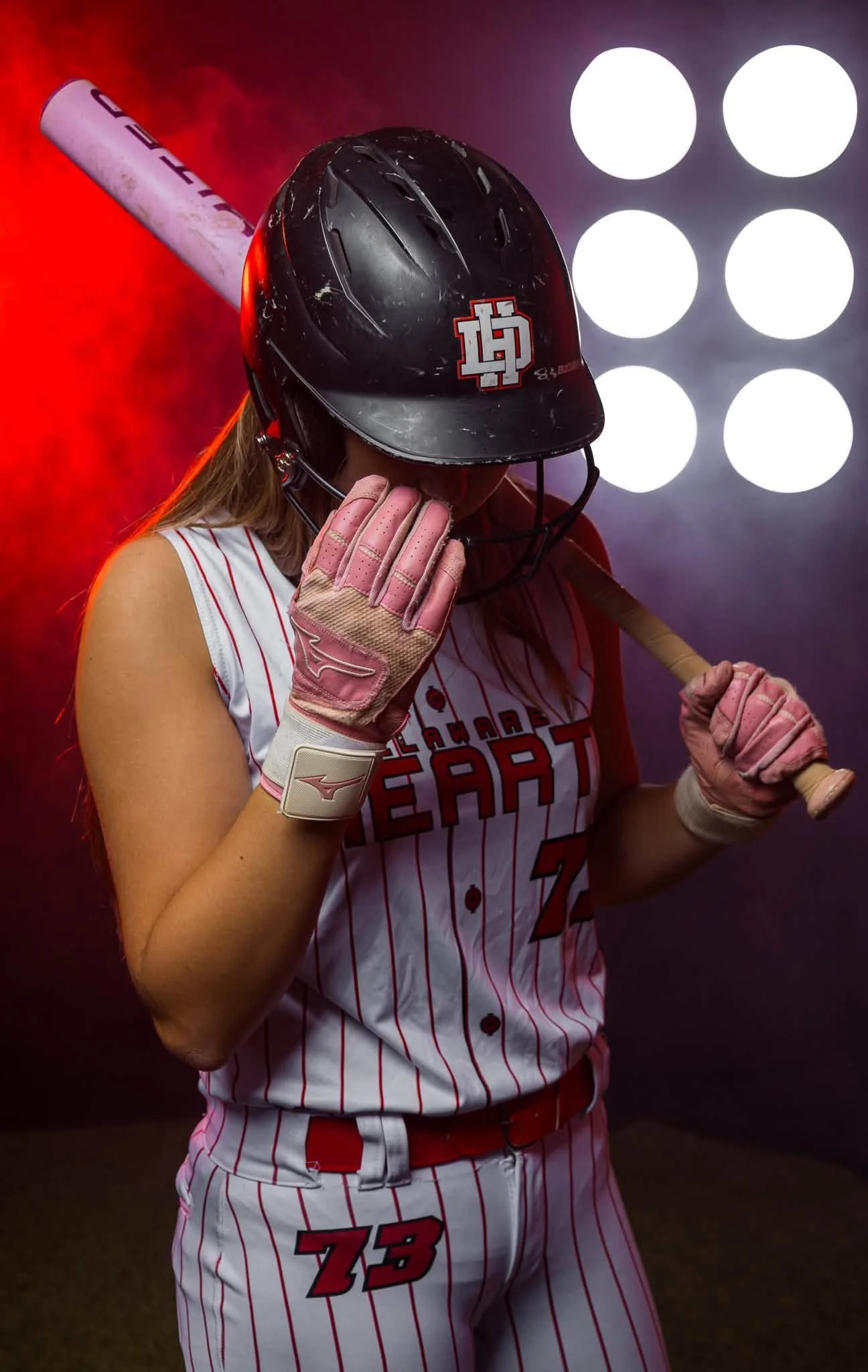 A female softball player wearing a black helmet with a red and white logo, pink gloves, and a white and red striped uniform with the number 78, holding a bat over her shoulder, standing in front of a dark background with bright white lights.