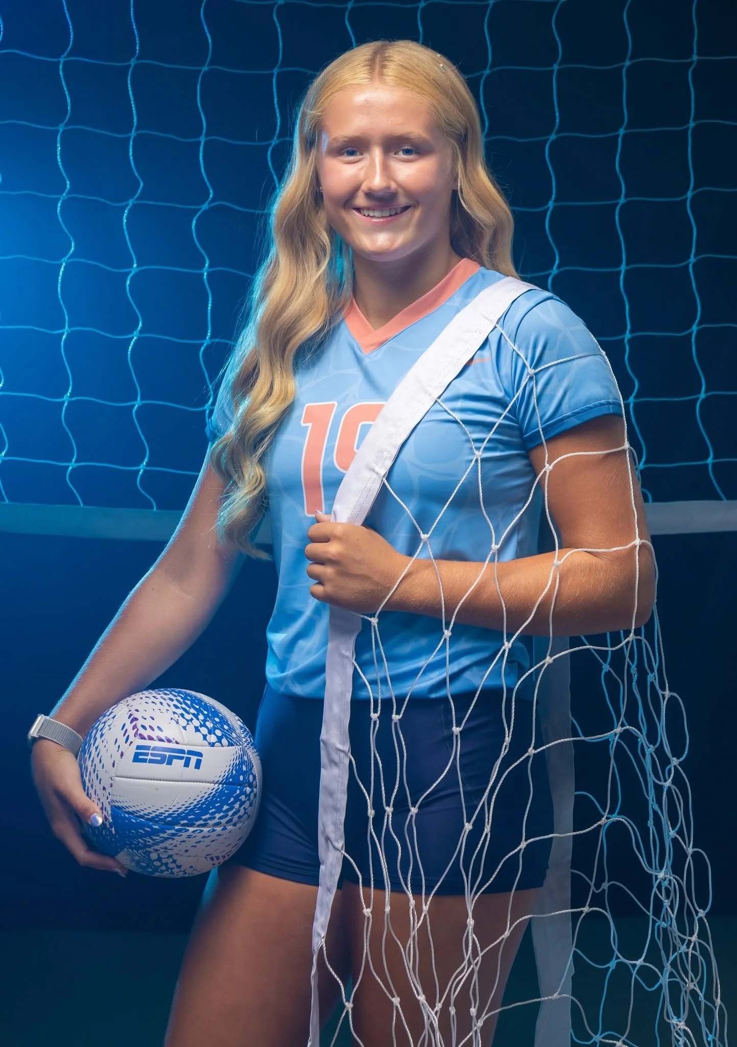 A young woman with long blonde hair, wearing a blue sports uniform with the number 10, standing behind a volleyball net with a volleyball in her hand, smiling at the camera, in a sports setting.