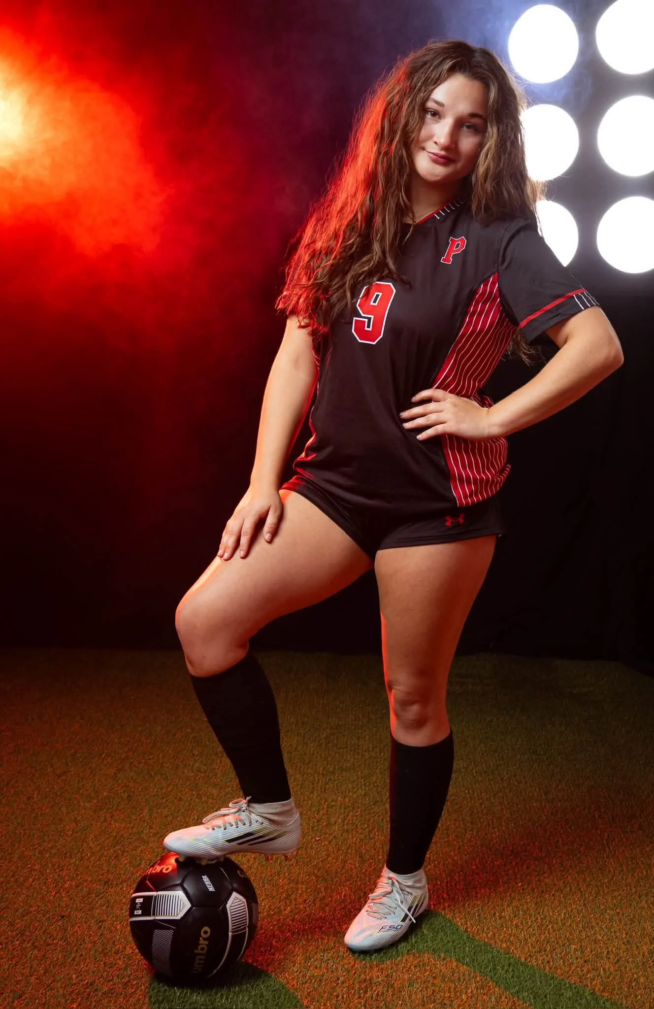 Young woman in soccer uniform standing with her hand on her hip, one foot on a soccer ball, in a studio with bright light background.