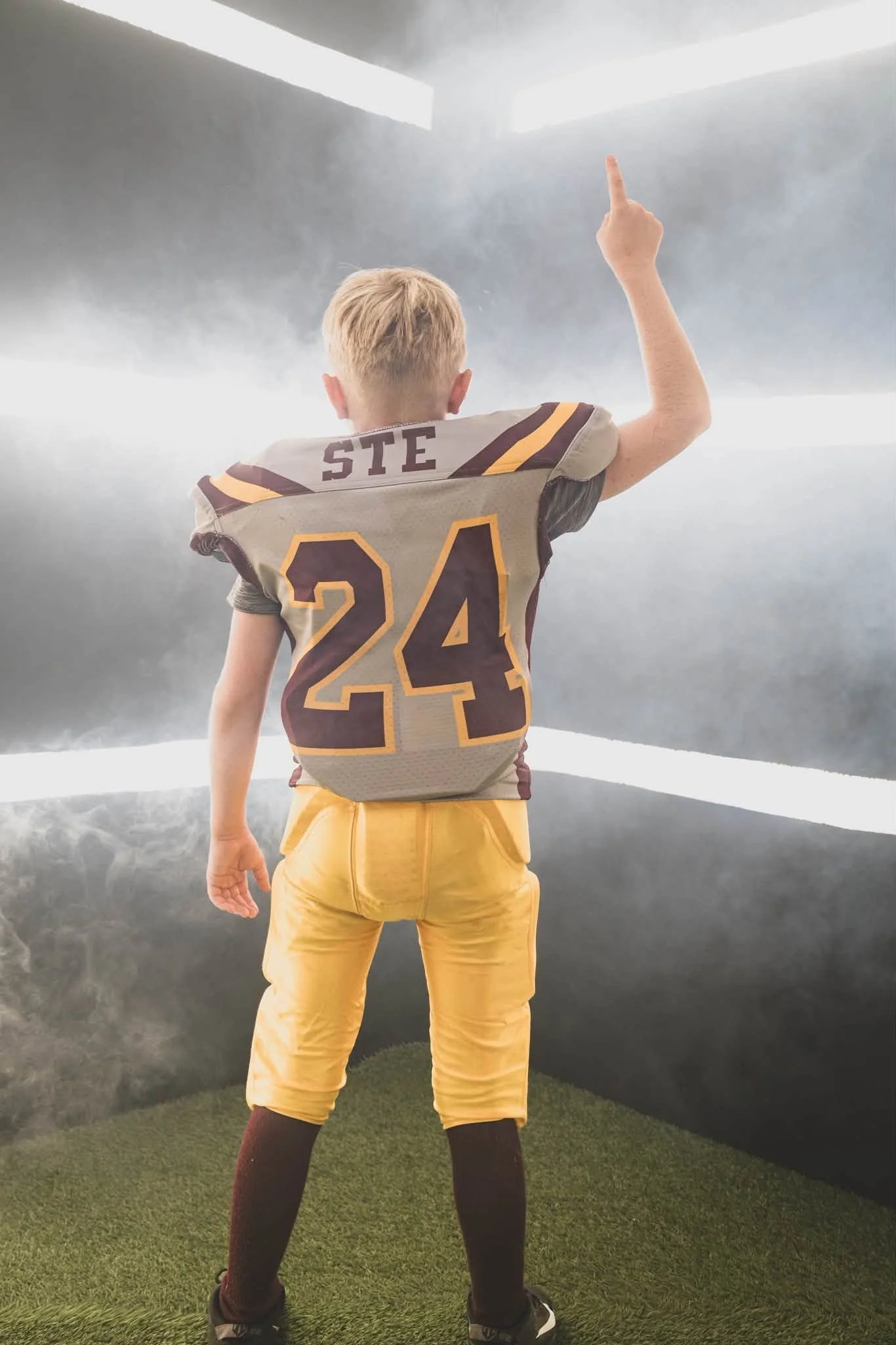 Back view of a young football player in uniform, raising his right hand with one finger pointing upwards, standing on artificial grass in a room with a foggy mountain landscape mural background.