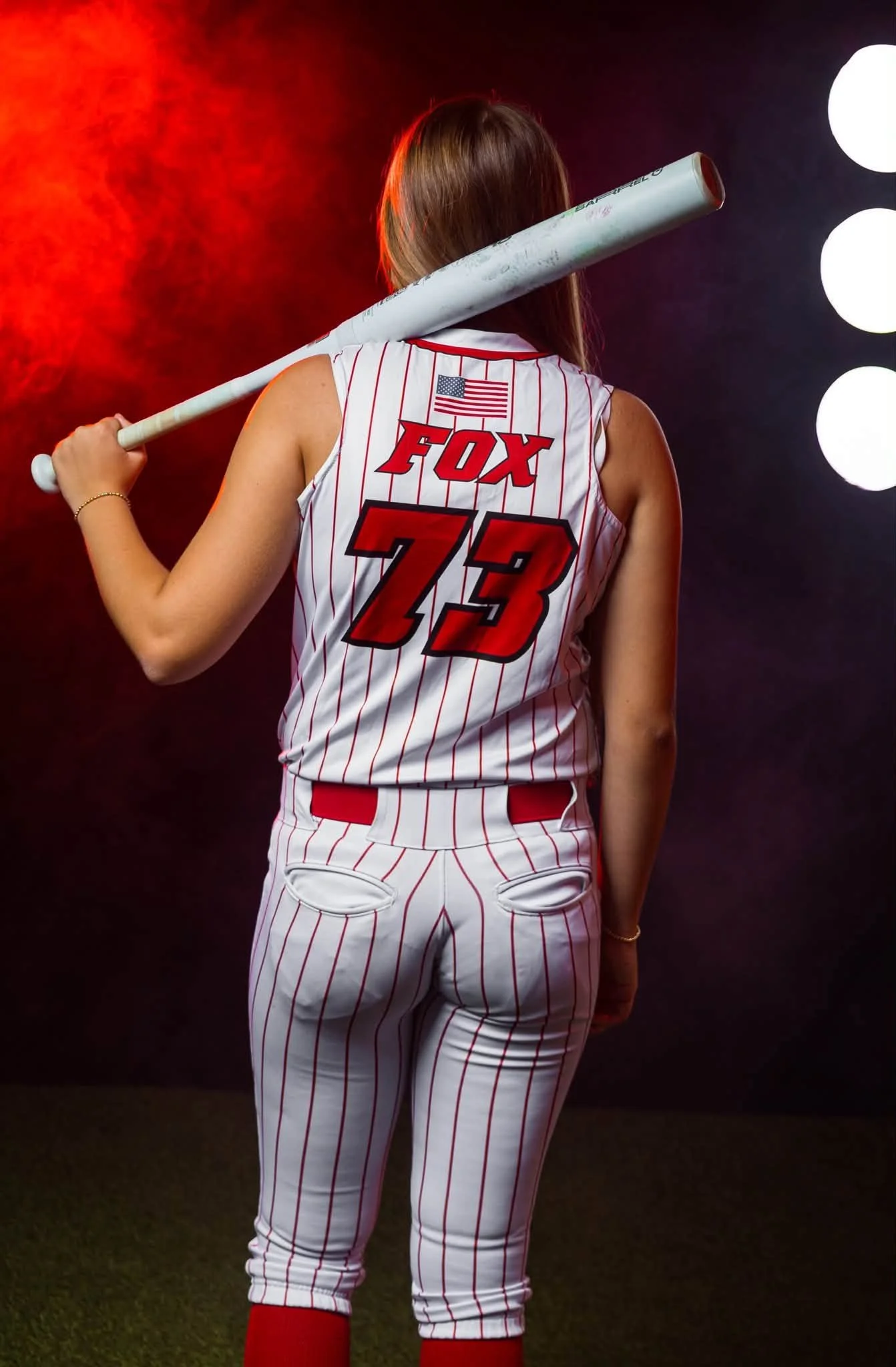 A woman in a baseball jersey with the name 'Fox' and the number 23 on the back, holding a baseball bat on her shoulder, standing against a dark background with bright lights.