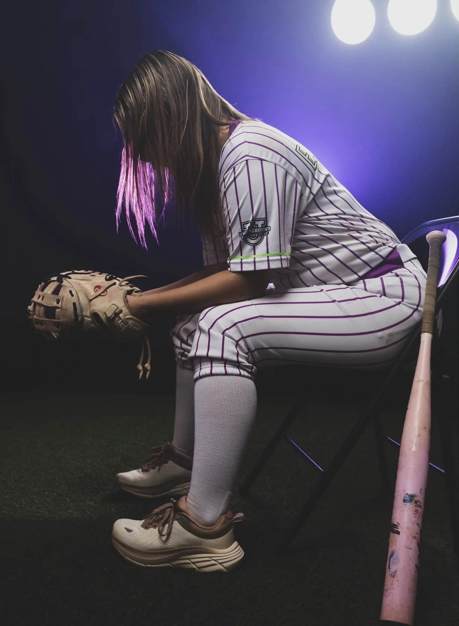 A girl dressed in baseball gear, including striped jersey, gray pants, socks, and sneakers, sits on a chair with a baseball glove in her hands, in a dimly lit room with purple lighting and fluorescent lights in the background.