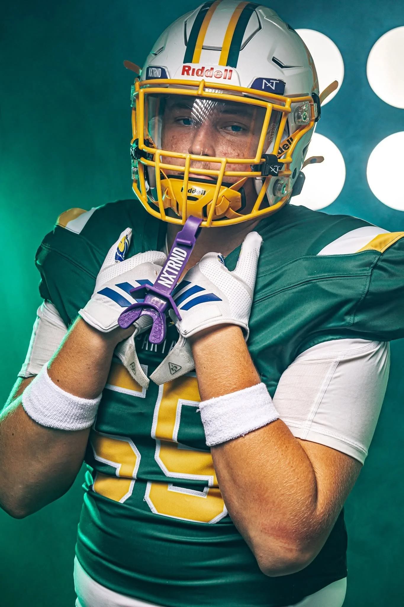 A football player wearing a green, yellow, and white uniform and a white helmet with yellow and blue stripes, holding a purple medal with the word 'NIXTRND' written on it.