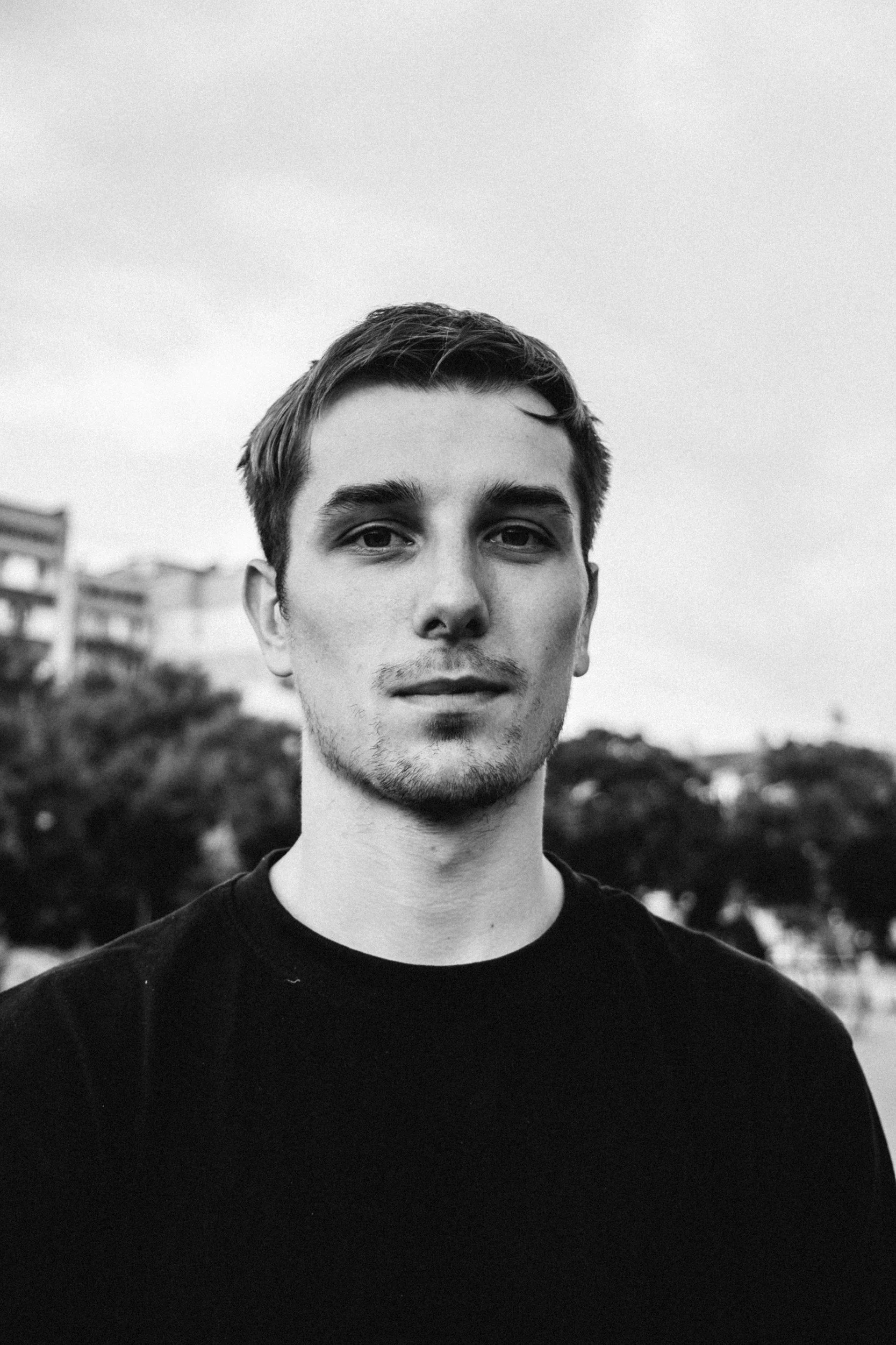 Black and white photo of a young man with short hair, wearing a black shirt, outdoors with trees and buildings in the background.