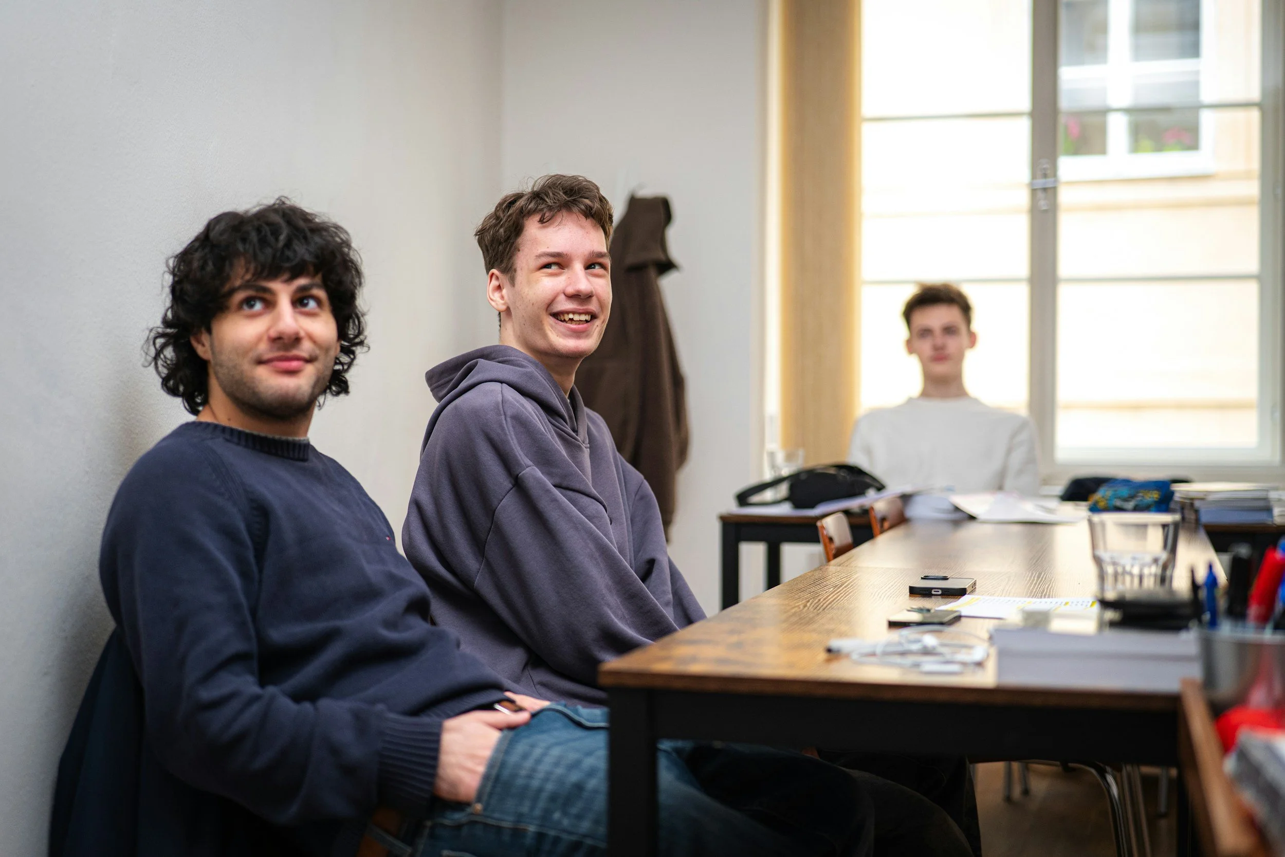 Three young men sitting at a wooden table in a room with a large window, with a fourth person in the background, smiling and looking at the camera.