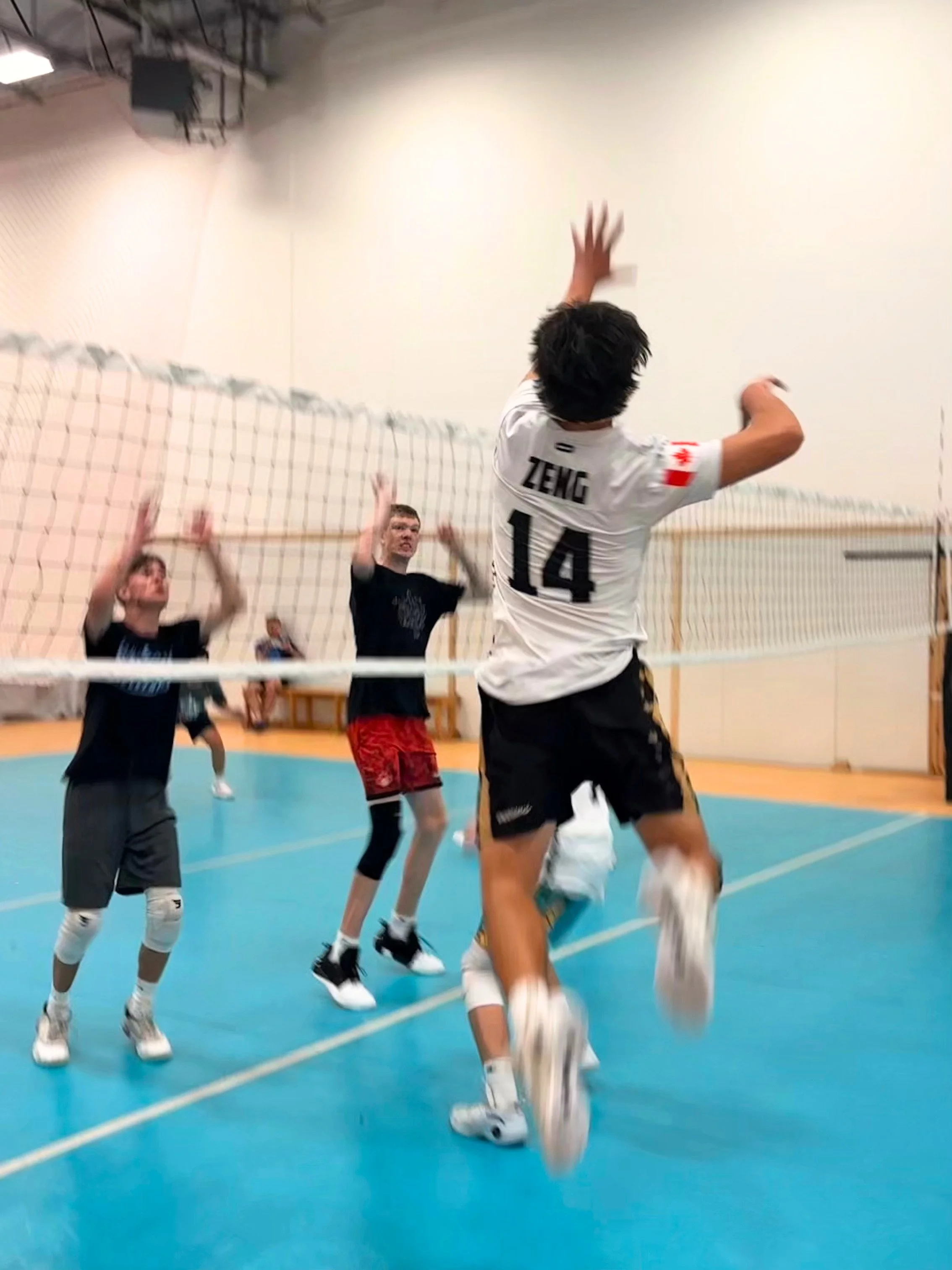 A group of young men playing volleyball indoors. One player, wearing a white jersey with the name 'Zeng' and number 14, is jumping to hit the ball over the net. Two other players are preparing to receive or defend, and a person in the background is watching. The court has blue flooring, and a volleyball net separates the teams.