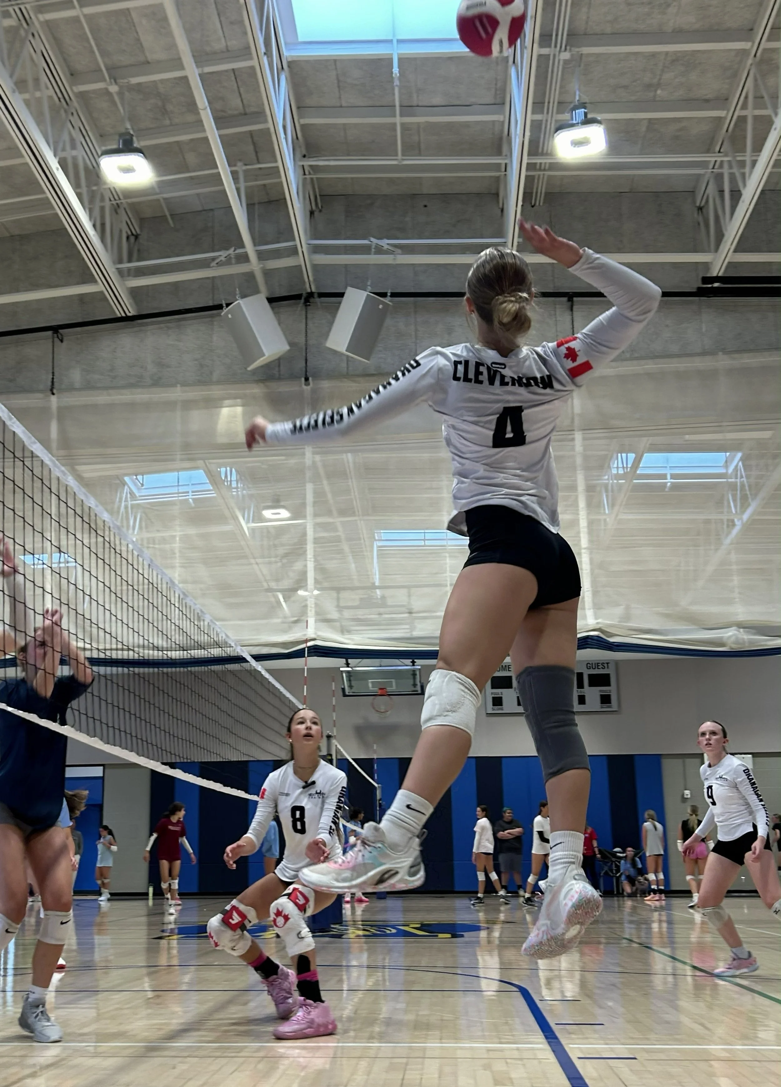 A female volleyball player in mid-air attempting to block or spike the ball during a game. She is wearing a white jersey with the name 'Clever' and the number 4. Other players and spectators are visible in the background inside an indoor sports gym.