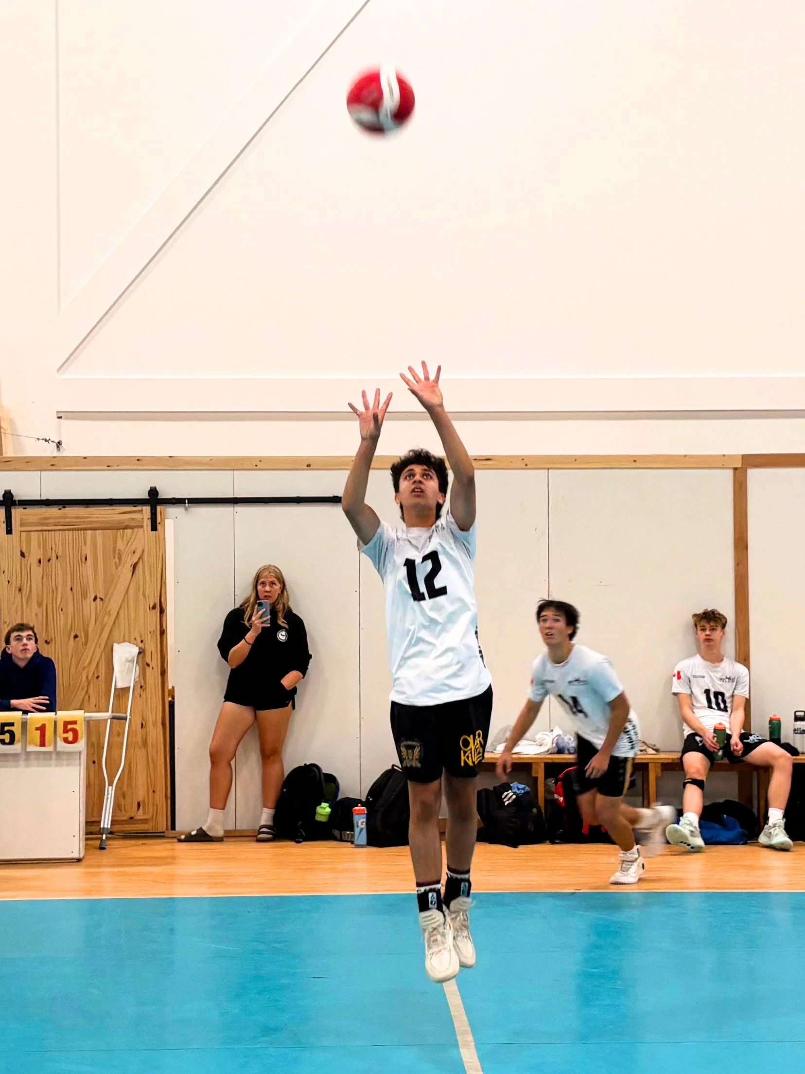 A volleyball player with the number 12 on his shirt is jumping to hit the volleyball during a game. Several other players, a woman taking a photo, and benches with water bottles are visible in the background.