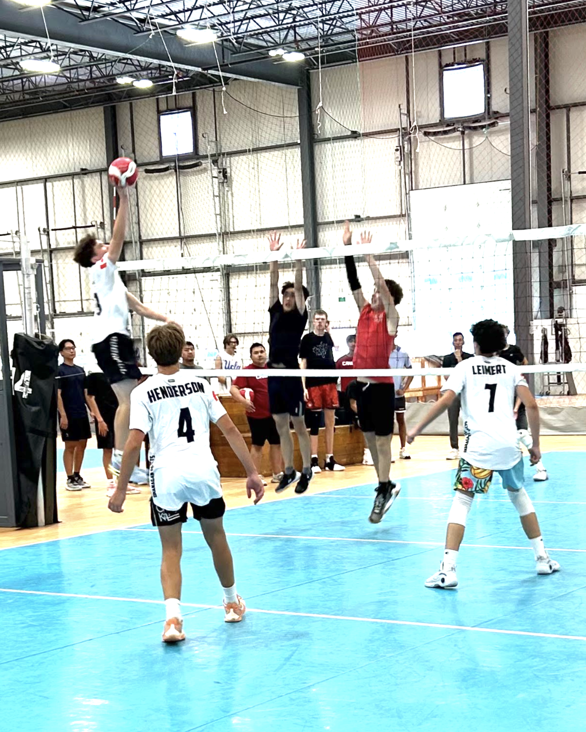 People playing indoor volleyball, with one player jumping to hit the ball over the net, while others attempt to block or receive.