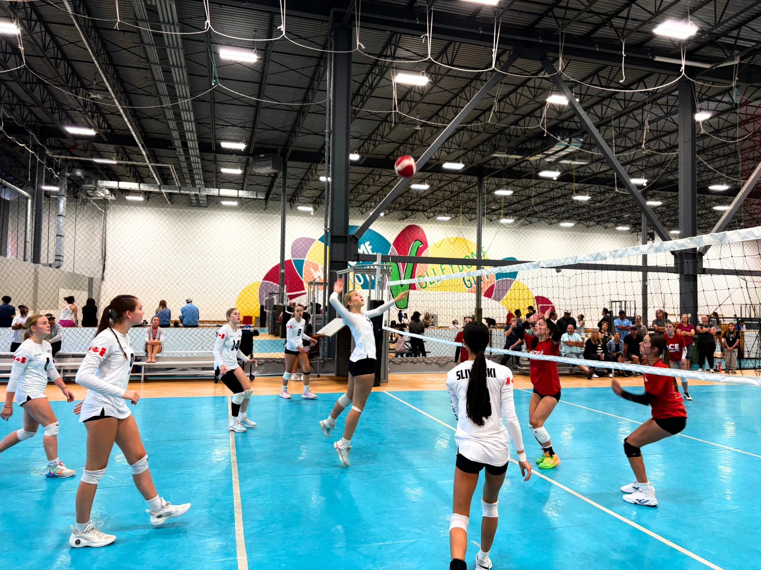 Indoor volleyball court with female players in white uniforms practicing, spectators seated along the sides, and a volleyball in mid-air near the net under a high ceiling with industrial lighting.