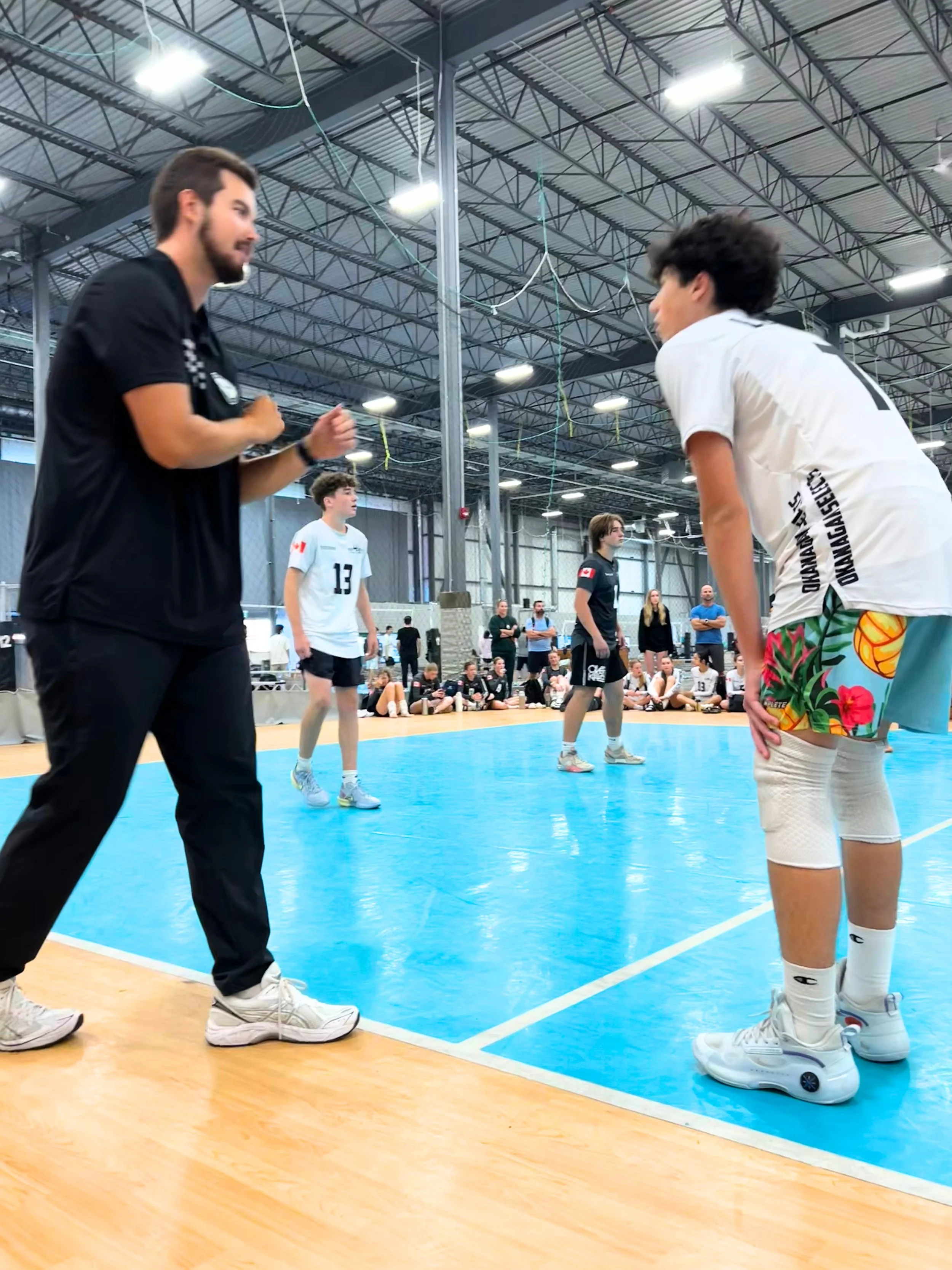A volleyball coach instructs players during a game in an indoor gymnasium. The players are wearing athletic gear, and the focus is on the girl wearing colorful shorts and roller skate sneakers. Spectators are seated in the background.