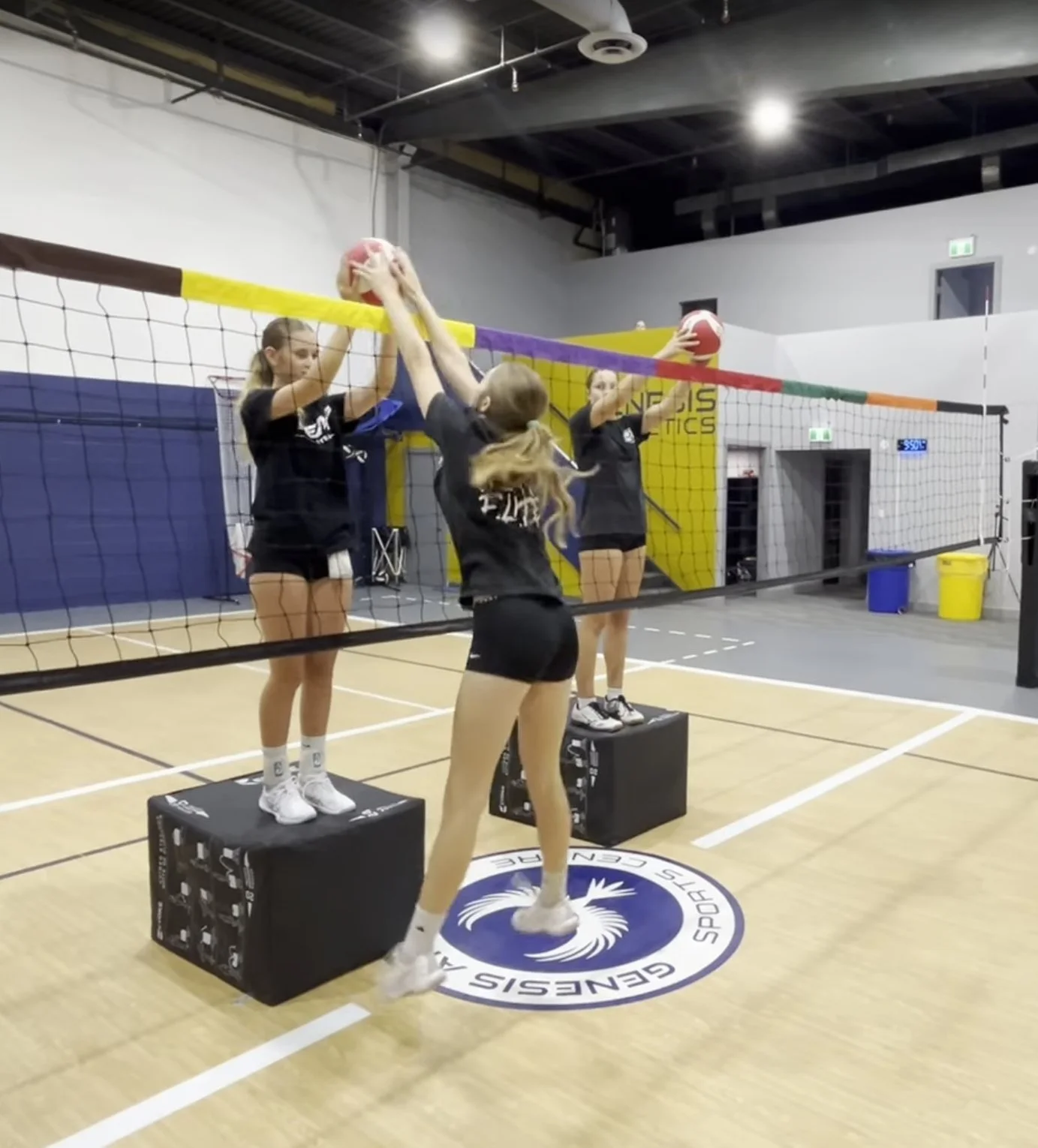 Three young female volleyball players practicing at an indoor gym, with two standing on blocks and one jumping in the air, hitting volleyballs over the net.