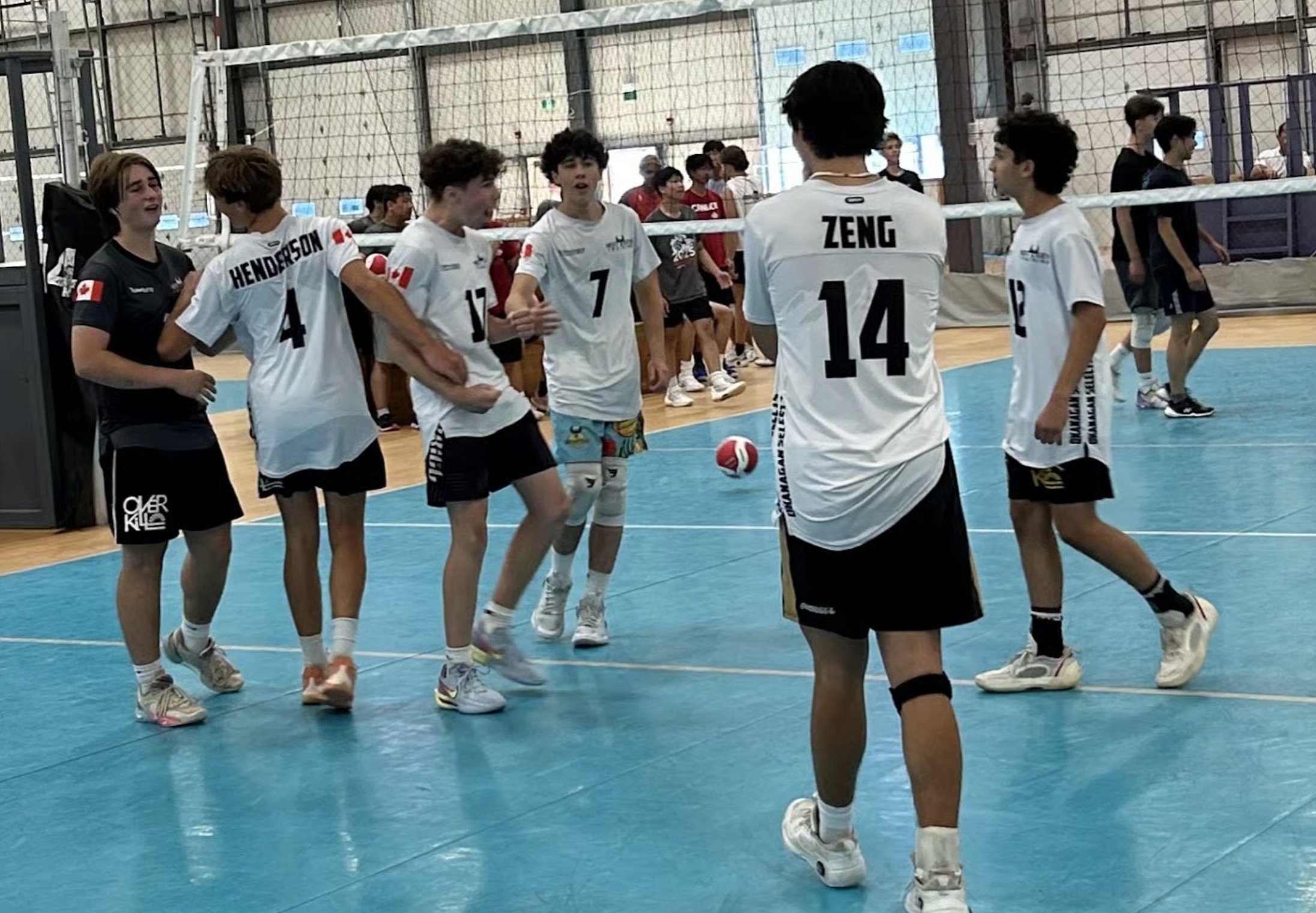 A group of young volleyball players on an indoor court, wearing white jerseys with black numbers and shorts, some with Canadian flags on their sleeves, standing near the net and engaging in conversation.